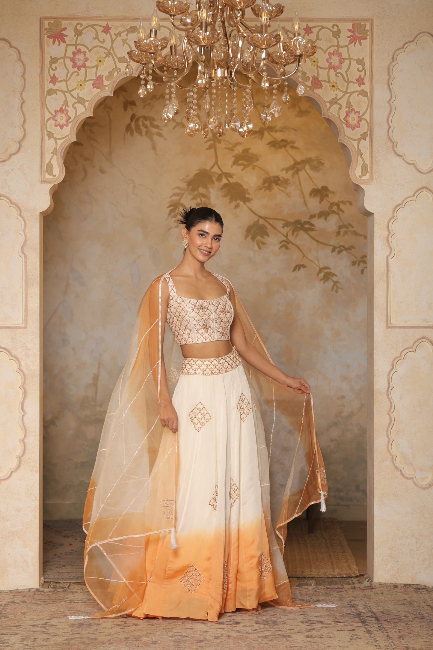 Woman in a traditional outfit with a decorative background. Sunanta Madaan – Ivory-Orange Satin Organza Lehenga, Embroidered Designer Outfit, Elegant Festive Attire, Premium Party & Reception Wear, Boho Indo-Western Dress, Indo-Western Fusion Wear, Indo-Western Attire for Engagement, Indo-Western Outfit for Sangeet, Indo-Western Draped & Embroidered Skirt, Mumbai, Delhi, Hyderabad, Bangalore, Pune, near me