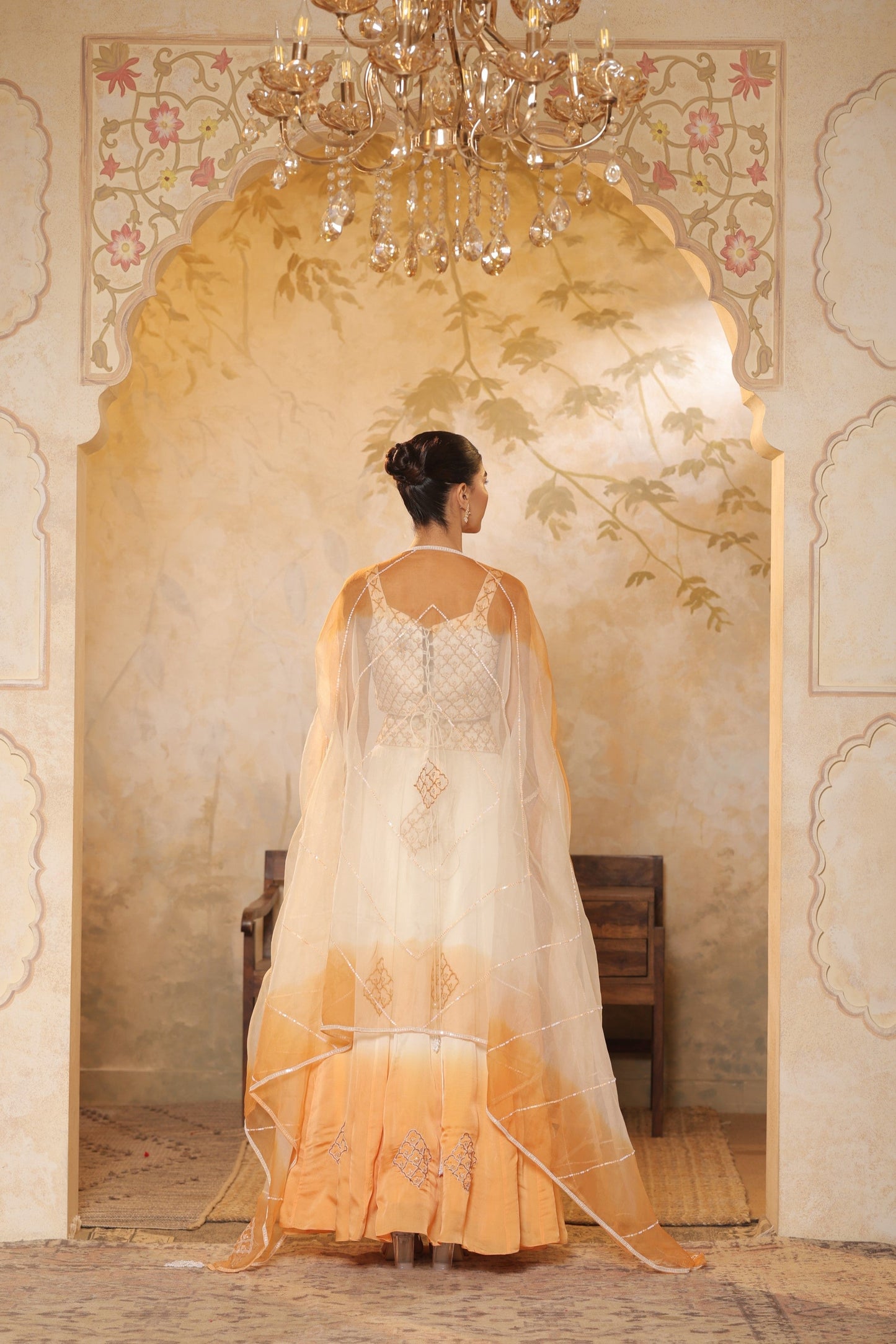 Woman in a traditional outfit standing in a decorated room with a chandelier. Sunanta Madaan – Ivory-Orange Satin Organza Lehenga, Embroidered Designer Outfit, Elegant Festive Attire, Premium Party & Reception Wear, Boho Indo-Western Dress, Indo-Western Fusion Wear, Indo-Western Attire for Engagement, Indo-Western Outfit for Sangeet, Indo-Western Draped & Embroidered Skirt, Mumbai, Delhi, Hyderabad, Bangalore, Pune, near me