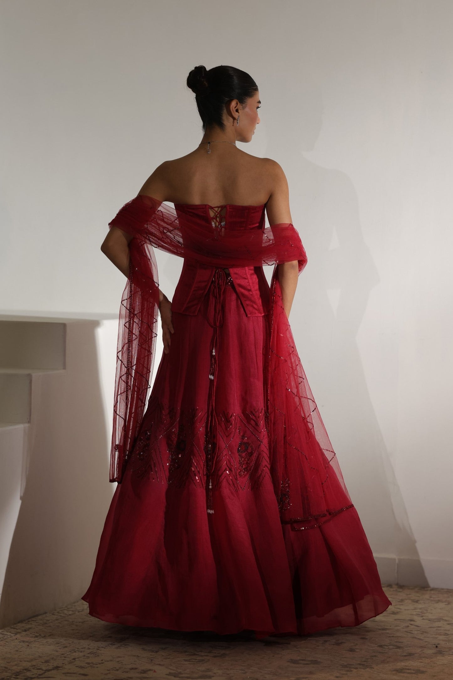 Woman wearing a red off-shoulder gown with sheer sleeves against a white wall. Sunanta Madaan – Burgundy Raw Silk & Organza Embroidered Lehenga, Elegant Bridal Wear, Premium Festive Outfit, Boho Indo-Western Dress, Indo-Western Fusion Wear, Indo-Western Attire for Engagement, Indo-Western Outfit for Sangeet, Indo-Western Draped & Embroidered Skirt, Women’s Designer Bridal Ensemble, Delhi, Jaipur, Hyderabad, Ahmedabad, Bangalore, near me