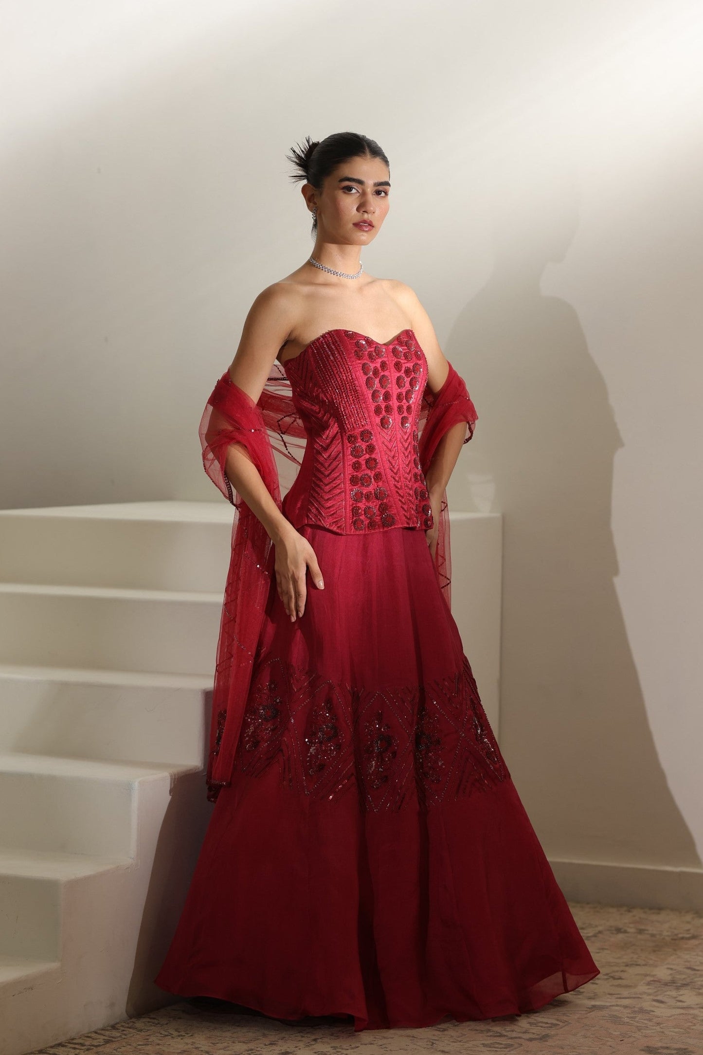 Woman in a red strapless gown standing on a staircase. Sunanta Madaan – Burgundy Raw Silk & Organza Embroidered Lehenga, Elegant Bridal Wear, Premium Festive Outfit, Boho Indo-Western Dress, Indo-Western Fusion Wear, Indo-Western Attire for Engagement, Indo-Western Outfit for Sangeet, Indo-Western Draped & Embroidered Skirt, Women’s Designer Bridal Ensemble, Delhi, Jaipur, Hyderabad, Ahmedabad, Bangalore, near me