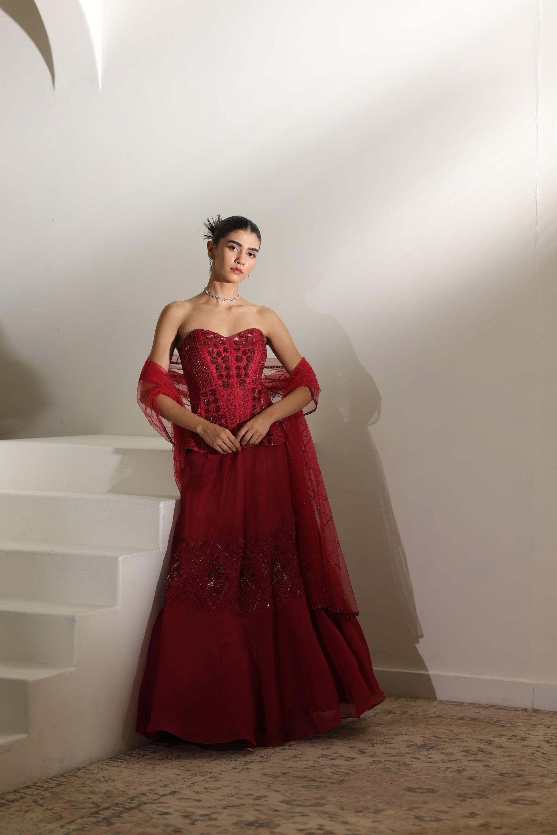 Woman in a red evening gown standing on a staircase; Sunanta Madaan – Burgundy Raw Silk & Organza Embroidered Lehenga, Elegant Bridal Wear, Premium Festive Outfit, Boho Indo-Western Dress, Indo-Western Fusion Wear, Indo-Western Attire for Engagement, Indo-Western Outfit for Sangeet, Indo-Western Draped & Embroidered Skirt, Women’s Designer Bridal Ensemble, Delhi, Jaipur, Hyderabad, Ahmedabad, Bangalore, near me