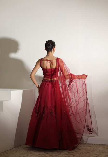 Woman in a red evening gown with a sheer overlay standing against a white wall. Sunanta Madaan – Burgundy Organza Hand-Embroidered Lehenga, Elegant Wedding Wear, Premium Festive Outfit, Boho Indo-Western Dress, Indo-Western Fusion Wear, Indo-Western Attire for Engagement, Indo-Western Outfit for Sangeet, Indo-Western Draped & Embroidered Skirt, Women’s Designer Bridal Ensemble, Mumbai, Jaipur, Hyderabad, Ahmedabad, Pune, near me