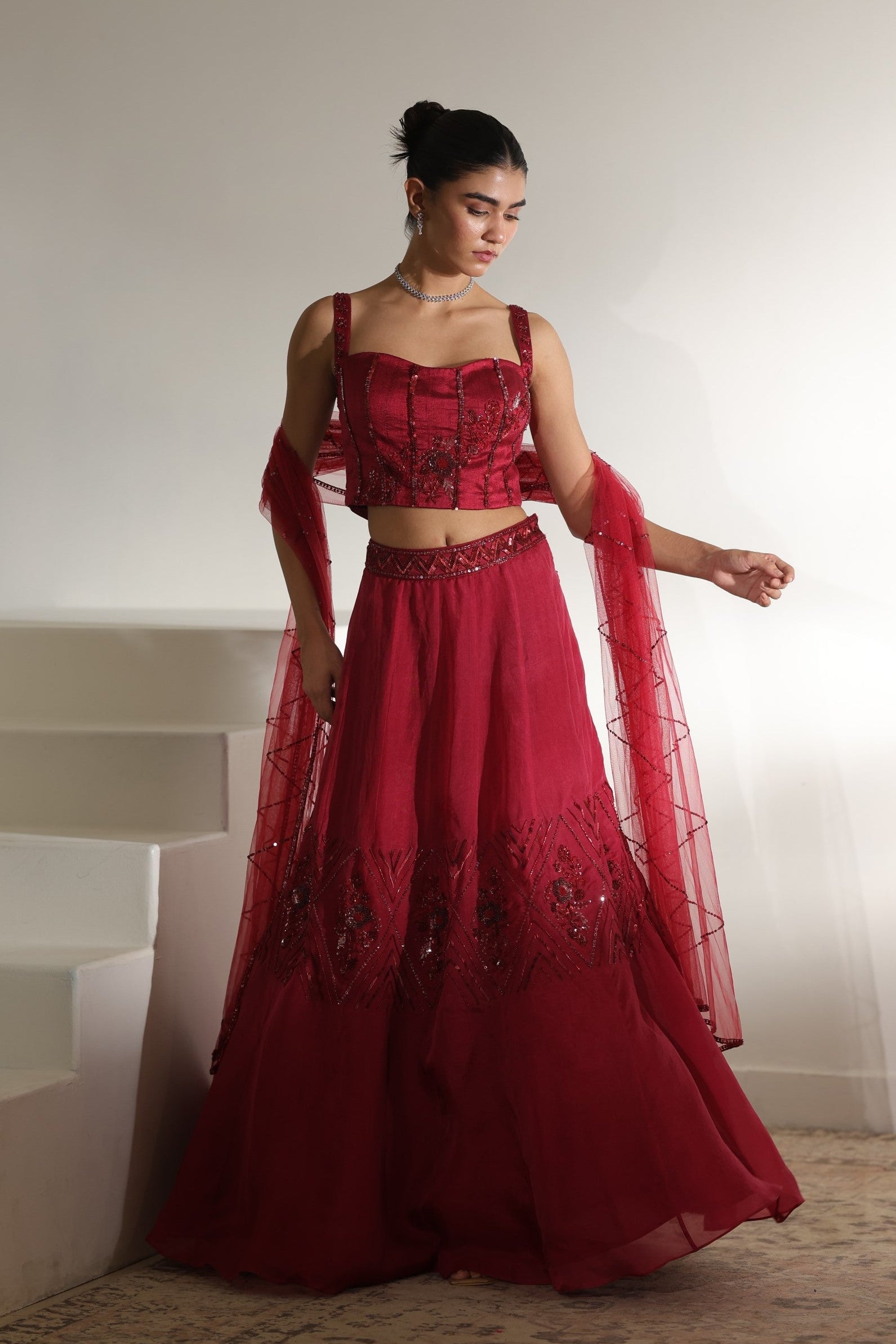Woman in a red traditional outfit standing against a white wall. Sunanta Madaan – Burgundy Organza Hand-Embroidered Lehenga, Elegant Wedding Wear, Premium Festive Outfit, Boho Indo-Western Dress, Indo-Western Fusion Wear, Indo-Western Attire for Engagement, Indo-Western Outfit for Sangeet, Indo-Western Draped & Embroidered Skirt, Women’s Designer Bridal Ensemble, Mumbai, Jaipur, Hyderabad, Ahmedabad, Pune, near me