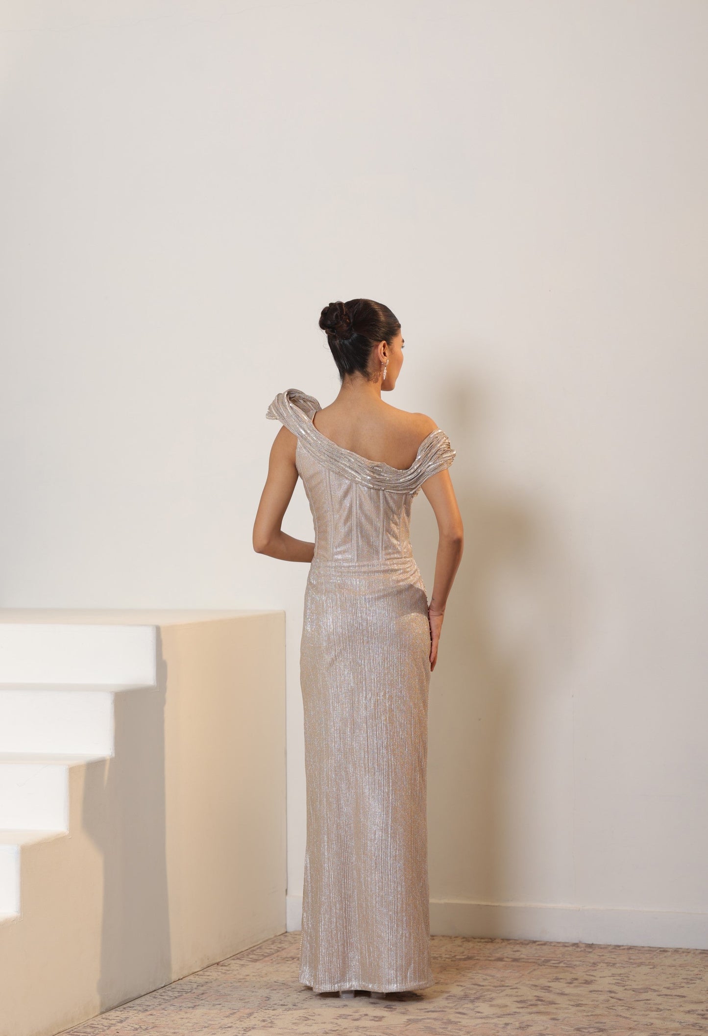 Woman in a silver off-shoulder gown standing against a white wall. Sunanta Madaan – Champagne Metallic Lycra Crepe Embroidered Gown, Elegant Cocktail Wear, Evening Party Dress, Boho Indo-Western Dress, Indo-Western Fusion Wear, Women’s Formal Gown, Indo-Western Outfit for Reception, Party Outfit, Indo-Western Draped Dress, Jaipur, Delhi, Pune, Ahmedabad, Hyderabad, near me