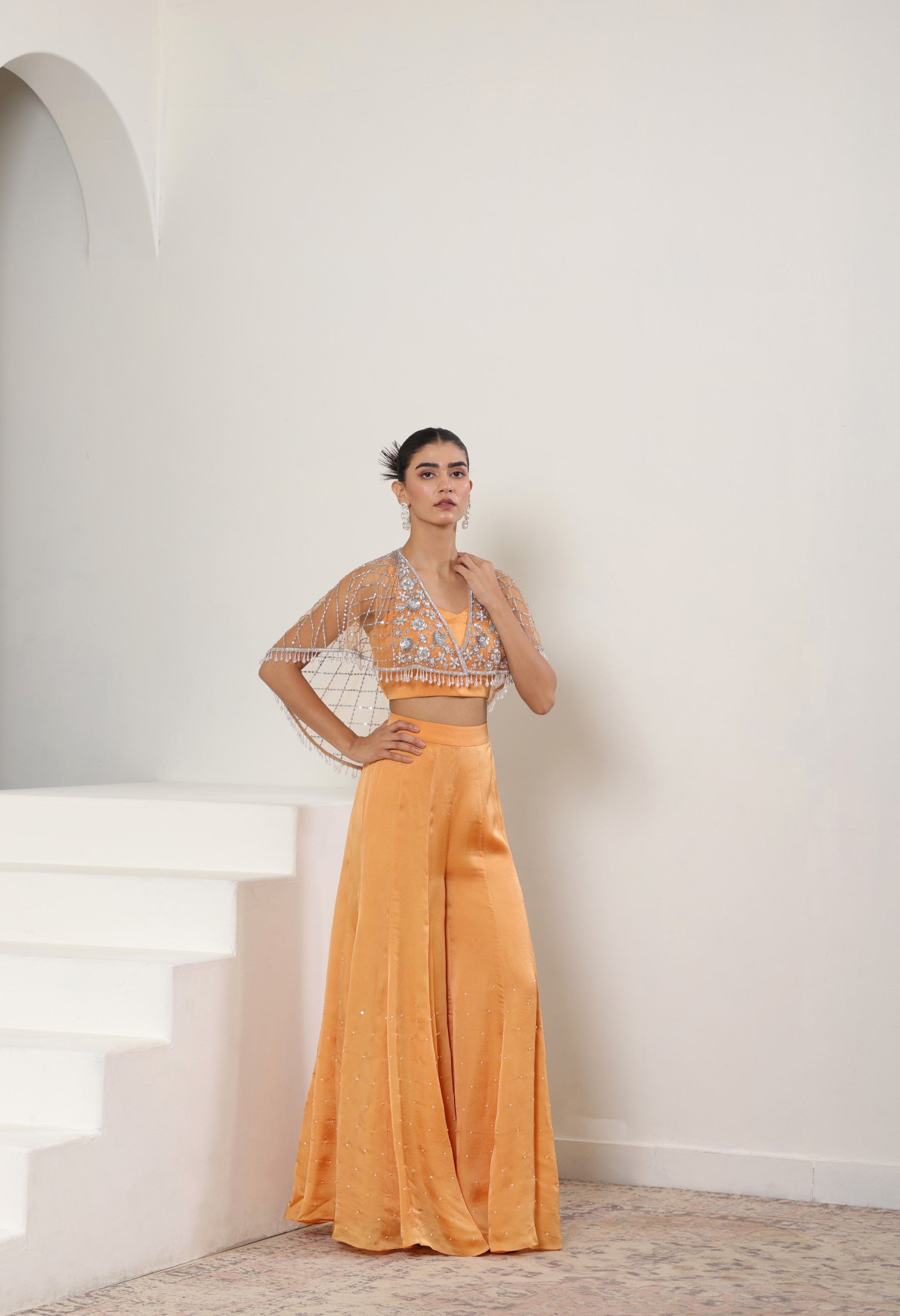 Woman wearing a yellow outfit with a white wall background; Orange satin organza sharara, embroidered cape set, festive ethnic outfit, elegant designer wear, premium traditional attire, Sunanta Madaan, near me, Mumbai, Delhi, Hyderabad, Bangalore, Pune