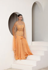 Woman in a traditional outfit standing on white steps with a white wall and archway in the background; Orange silk crepe sharara, draped festive outfit, elegant ethnic wear, designer traditional attire, premium party ensemble, Sunanta Madaan, near me, Mumbai, Delhi, Hyderabad, Bangalore, Pune