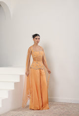 Woman wearing an orange evening gown with intricate detailing in a white room. Orange silk crepe sharara, draped festive outfit, elegant ethnic wear, designer traditional attire, premium party ensemble, Sunanta Madaan, near me, Mumbai, Delhi, Hyderabad, Bangalore, Pune