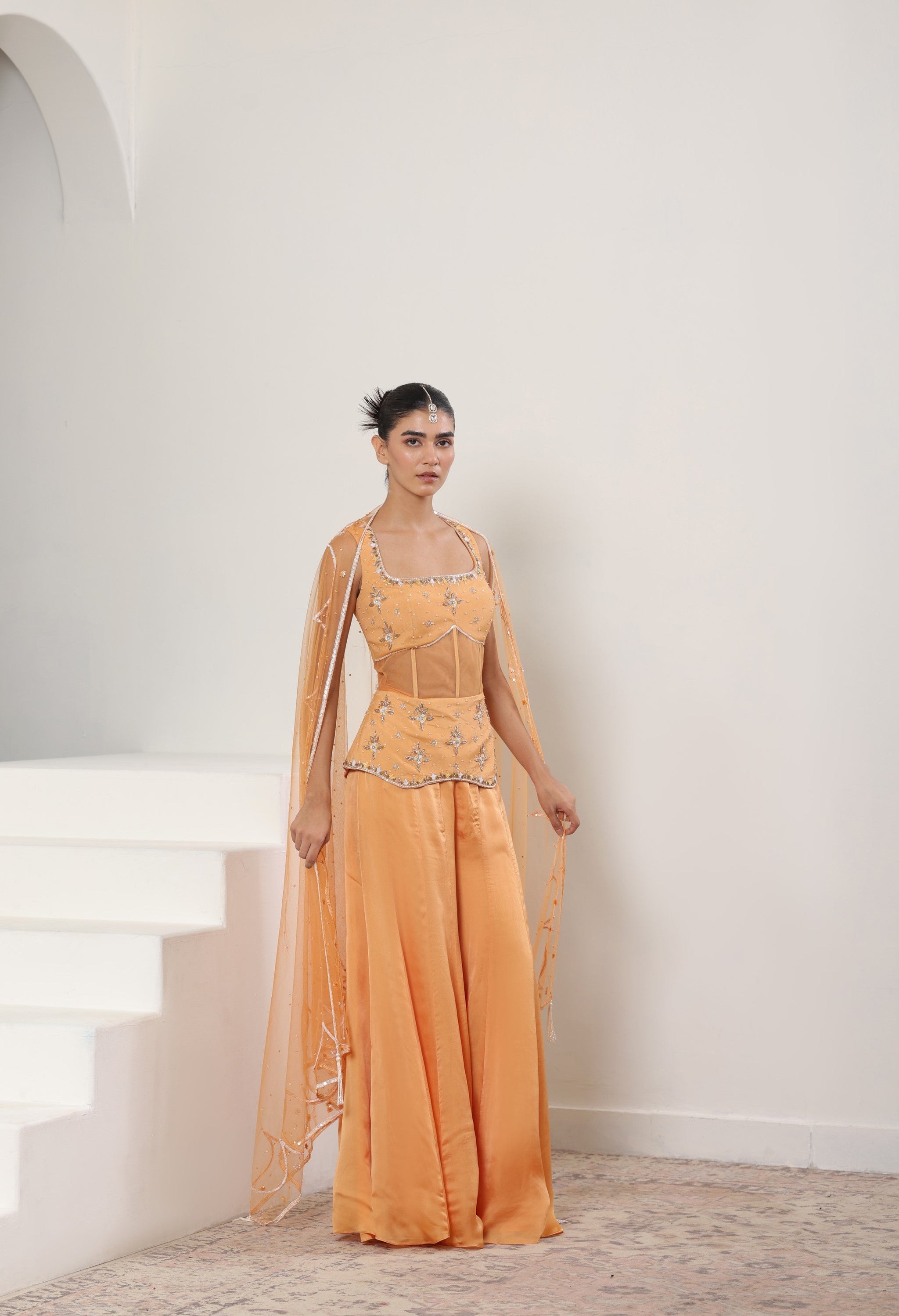 Woman wearing an orange evening gown with intricate detailing in a white room. Orange silk crepe sharara, draped festive outfit, elegant ethnic wear, designer traditional attire, premium party ensemble, Sunanta Madaan, near me, Mumbai, Delhi, Hyderabad, Bangalore, Pune