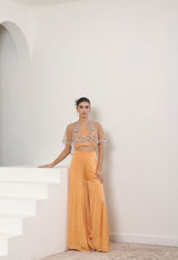 Woman in a peach outfit with intricate top and flowing pants standing on a white staircase. Orange satin organza sharara, embroidered cape set, festive ethnic outfit, elegant designer wear, premium traditional attire, Sunanta Madaan, near me, Mumbai, Delhi, Hyderabad, Bangalore, Pune