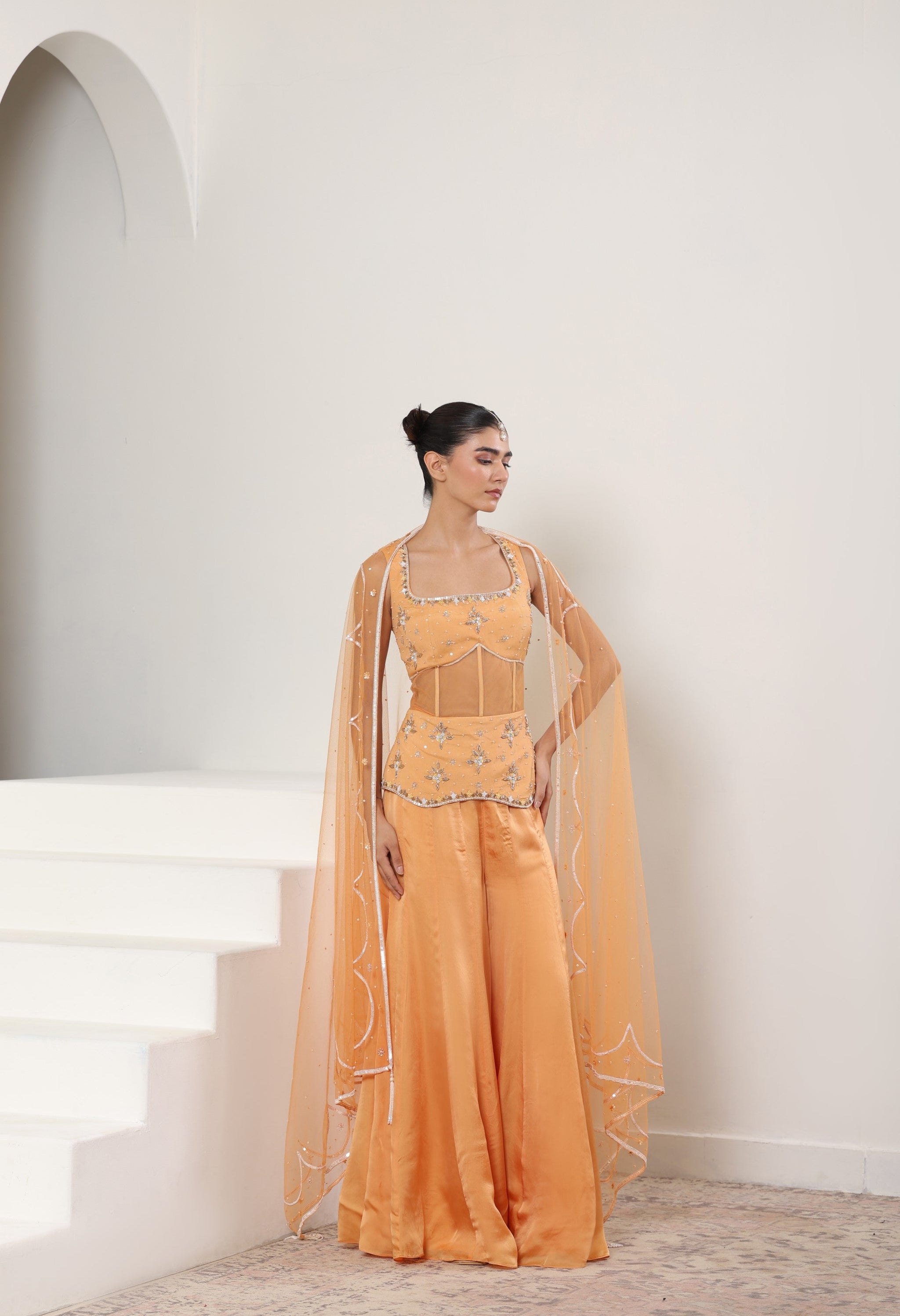 Woman in a peach-colored traditional outfit standing on a staircase with a white wall background; Orange silk crepe sharara, draped festive outfit, elegant ethnic wear, designer traditional attire, premium party ensemble, Sunanta Madaan, near me, Mumbai, Delhi, Hyderabad, Bangalore, Pune