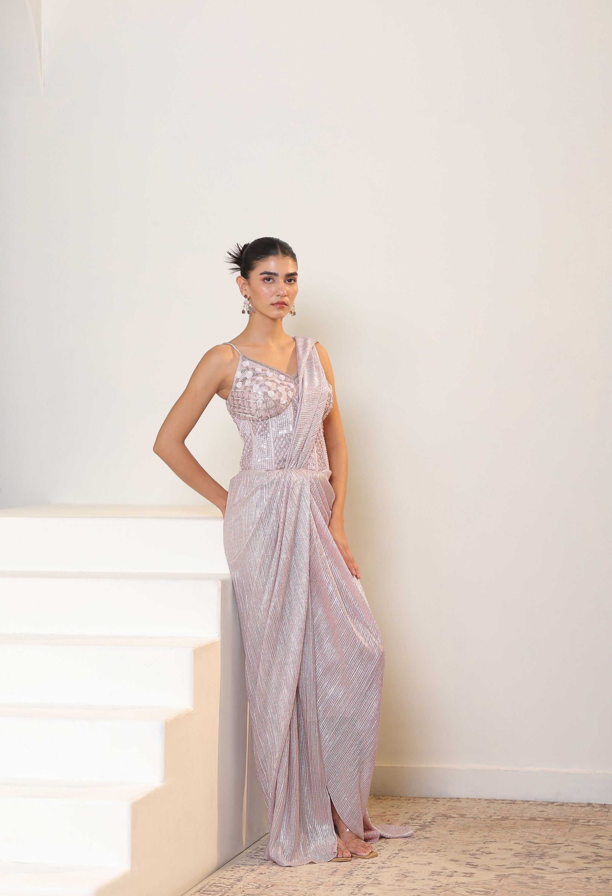 Woman in a silver evening gown standing on a staircase with a white wall background; Sunanta Madaan – Pink Metallic Lycra Crepe Saree, Premium Reception Wear, Elegant Party Outfit, Boho Indo-Western Dress, Indo-Western Fusion Wear, Women’s Formal Evening Dress, Indo-Western Outfit for Reception, Party Outfit, Indo-Western Draped & Embroidered Saree, Delhi, Jaipur, Hyderabad, Ahmedabad, Bangalore, near me