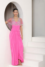 Woman wearing a pink dress with floral embellishments in a white architectural setting; Pink crepe skirt set, embroidered cape outfit, handcrafted engagement wear, elegant festive ensemble, designer ethnic attire, Sunanta Madaan