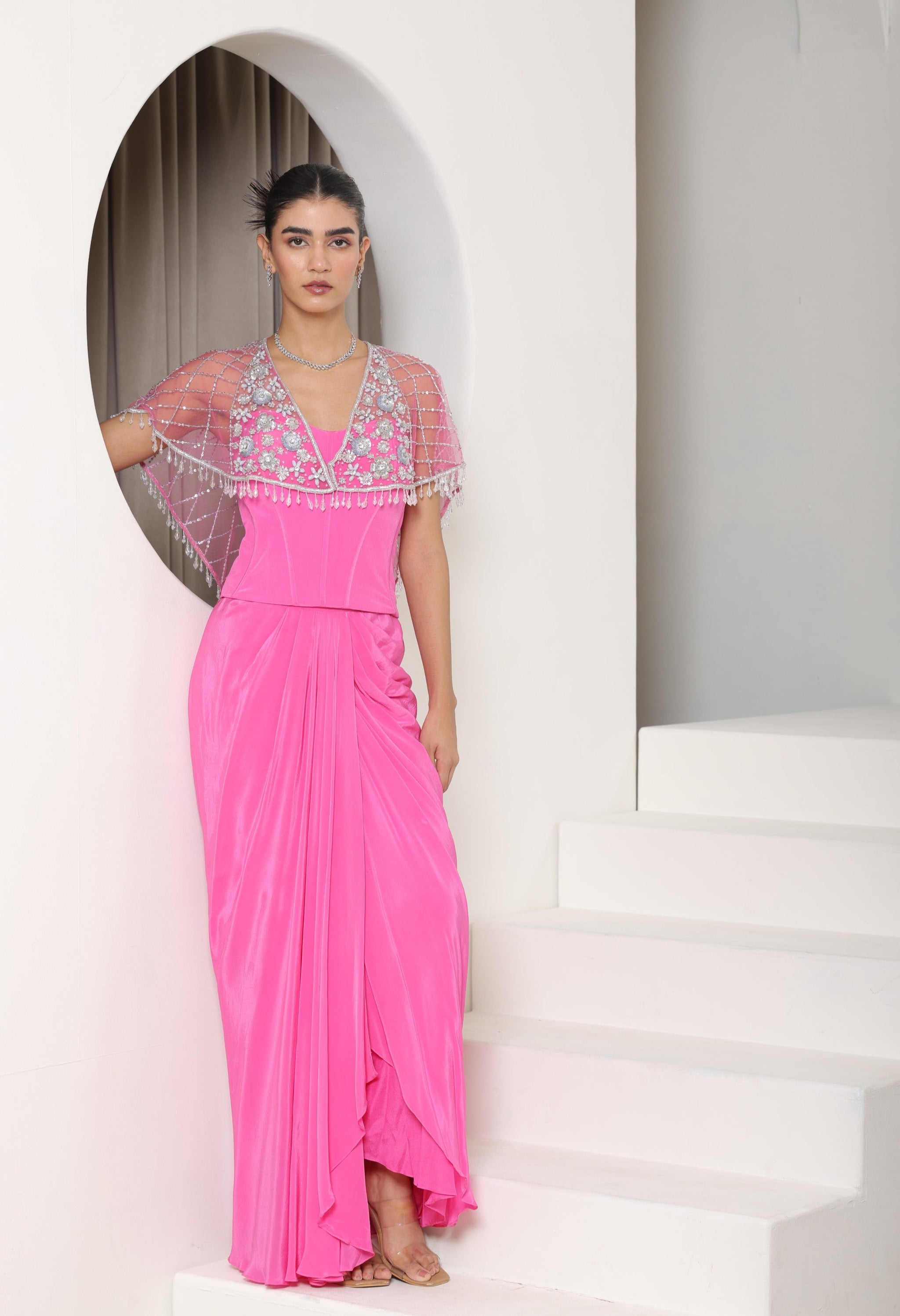 Woman wearing a pink dress with floral embellishments in a white architectural setting; Pink crepe skirt set, embroidered cape outfit, handcrafted engagement wear, elegant festive ensemble, designer ethnic attire, Sunanta Madaan