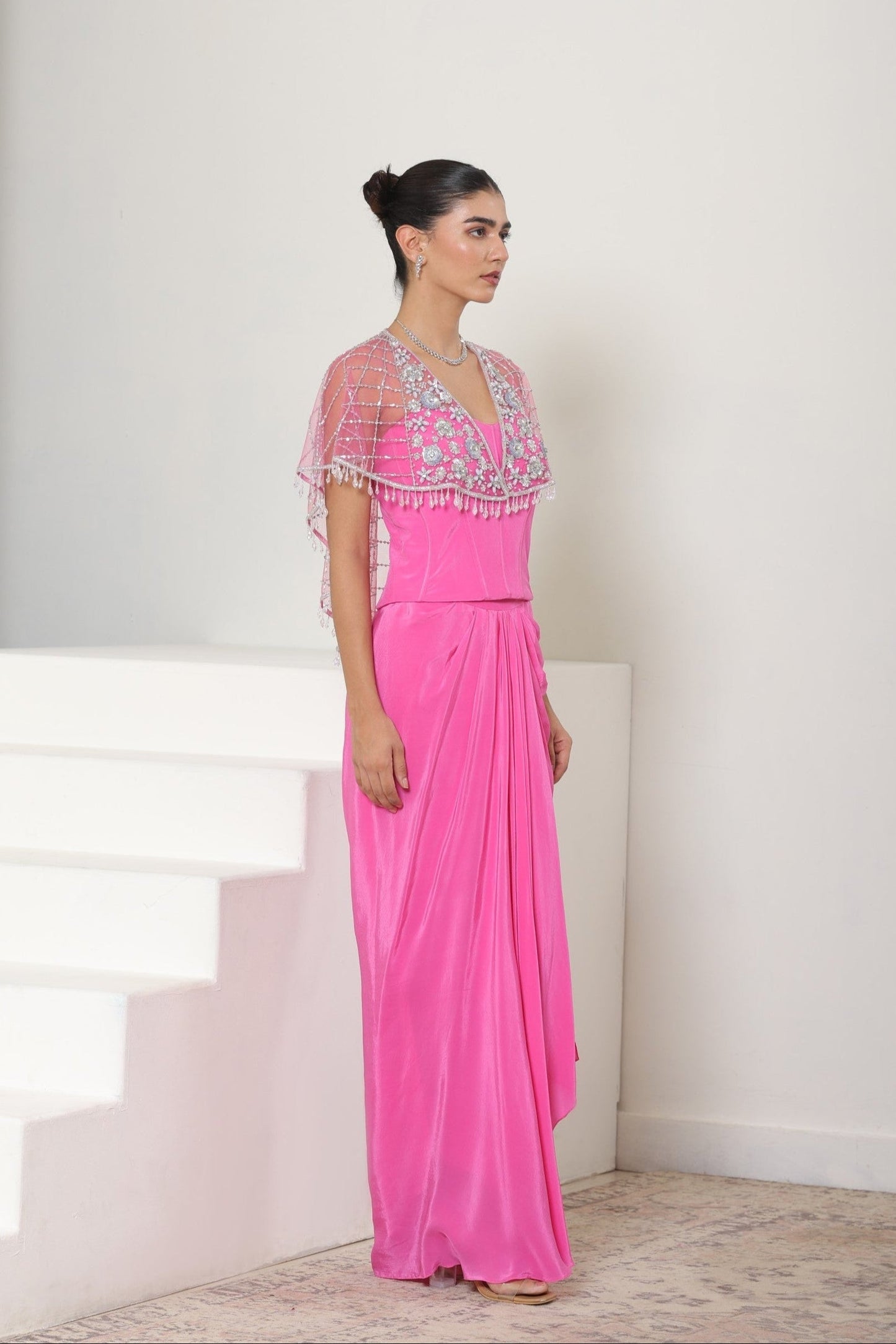Woman in a pink evening gown with a white background; Pink crepe skirt set, embroidered cape outfit, handcrafted engagement wear, elegant festive ensemble, designer ethnic attire, Sunanta Madaan