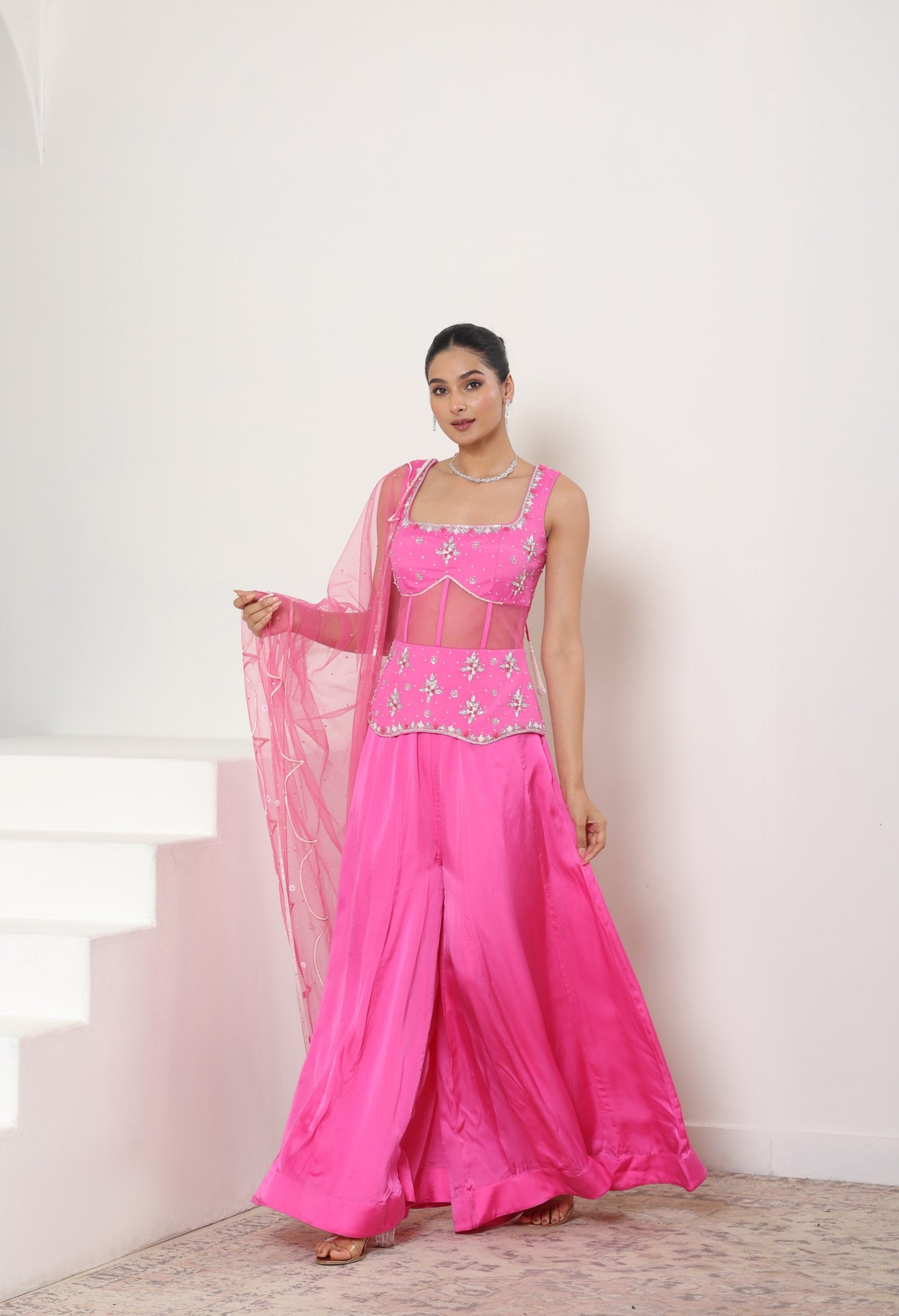 Woman in a pink traditional outfit standing on a staircase with a white wall background; Pink satin organza sharara, embroidered festive outfit, elegant occasional wear, designer ethnic attire, premium traditional set, Sunanta Madaan, near me, Mumbai, Delhi, Hyderabad, Bangalore, Pune