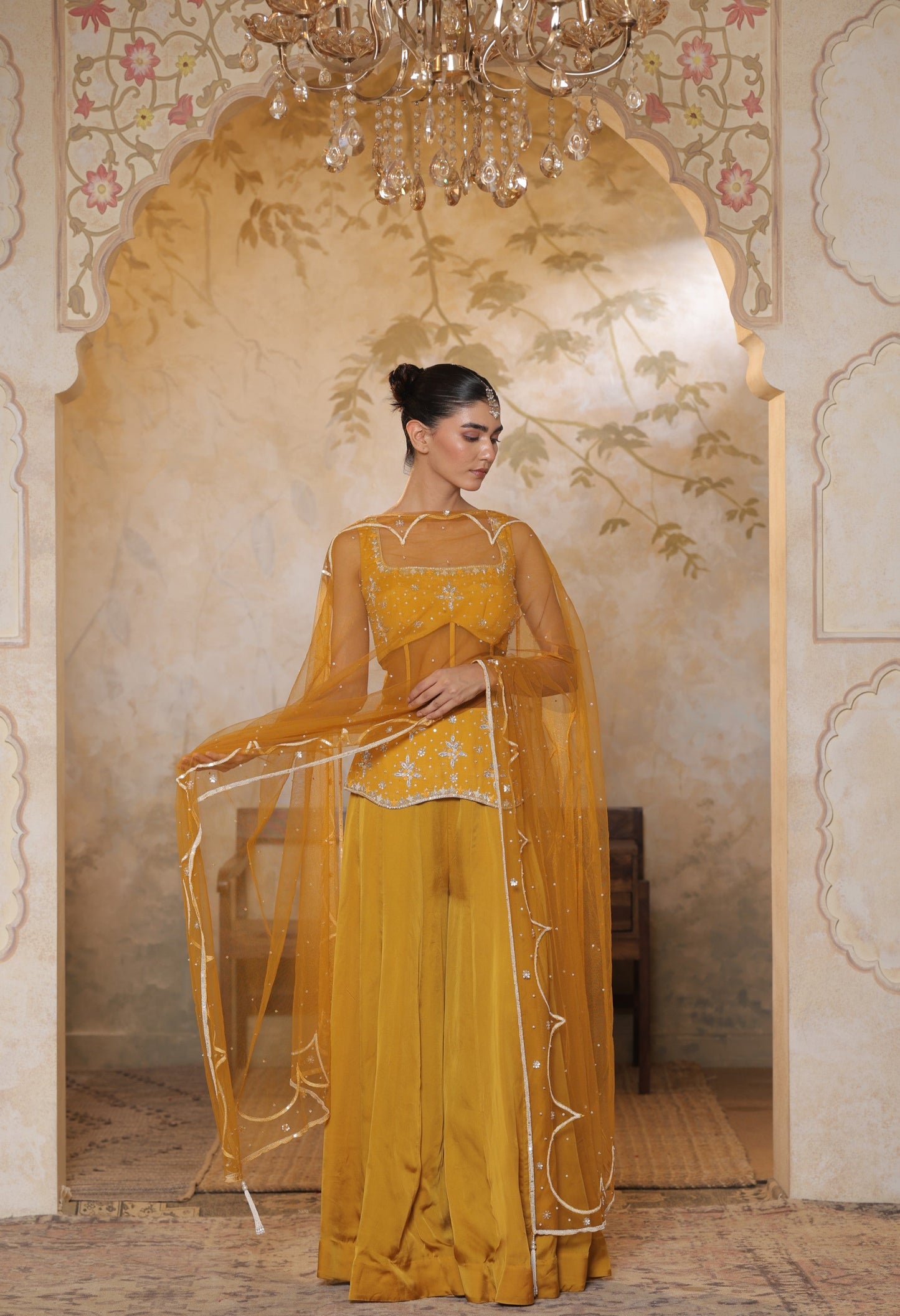 Woman in a yellow traditional outfit standing in an ornate room with a chandelier. Yellow silk crepe sharara, festive ethnic outfit, elegant occasional wear, designer traditional attire, premium party ensemble, Sunanta Madaan, near me, Mumbai, Delhi, Hyderabad, Bangalore, Pune