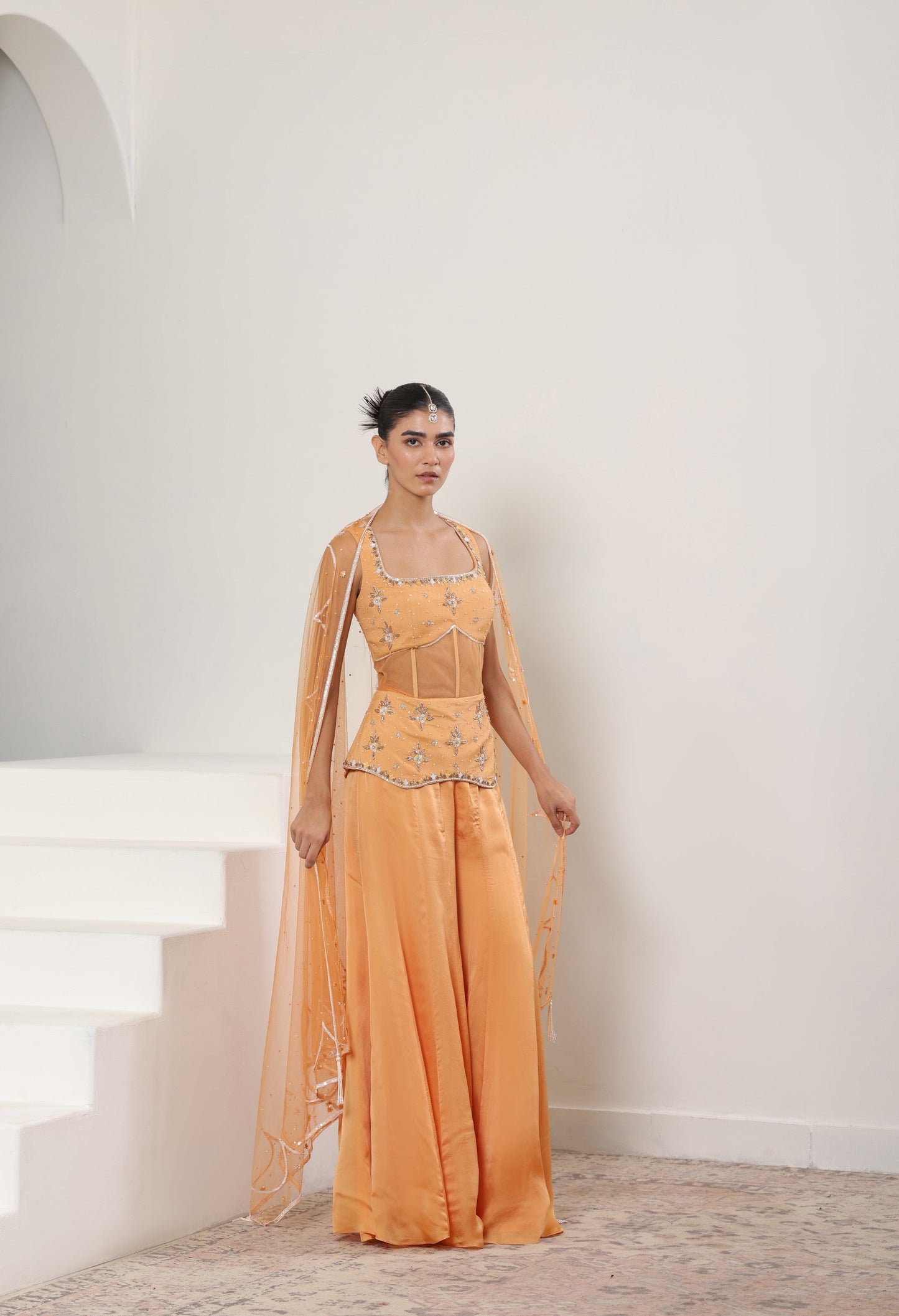Woman wearing an orange evening gown with intricate detailing in a white room. Orange silk crepe sharara, draped festive outfit, elegant ethnic wear, designer traditional attire, premium party ensemble, Sunanta Madaan, near me, Mumbai, Delhi, Hyderabad, Bangalore, Pune