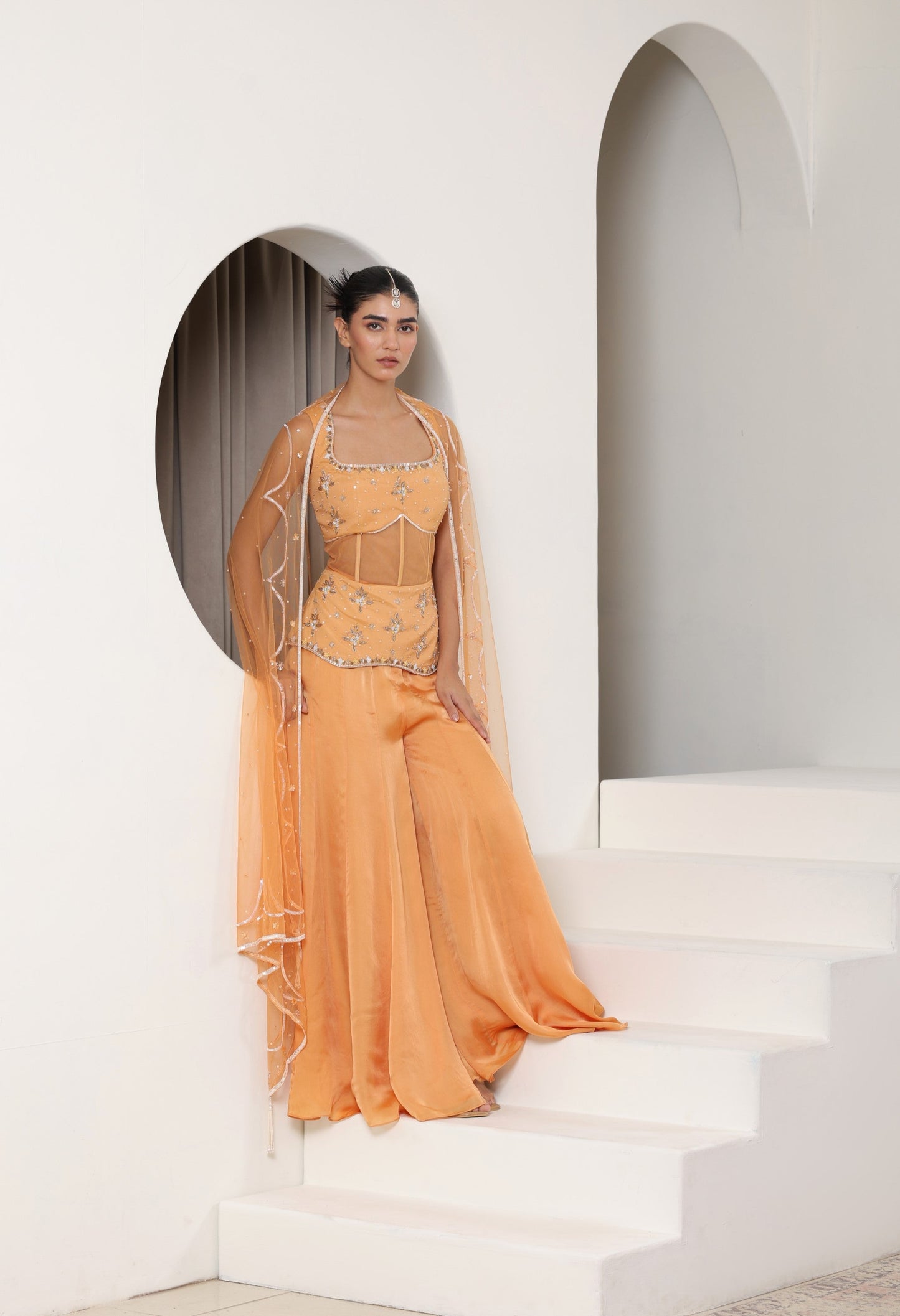 Woman in a traditional outfit standing on white steps with a white wall and archway in the background; Orange silk crepe sharara, draped festive outfit, elegant ethnic wear, designer traditional attire, premium party ensemble, Sunanta Madaan, near me, Mumbai, Delhi, Hyderabad, Bangalore, Pune