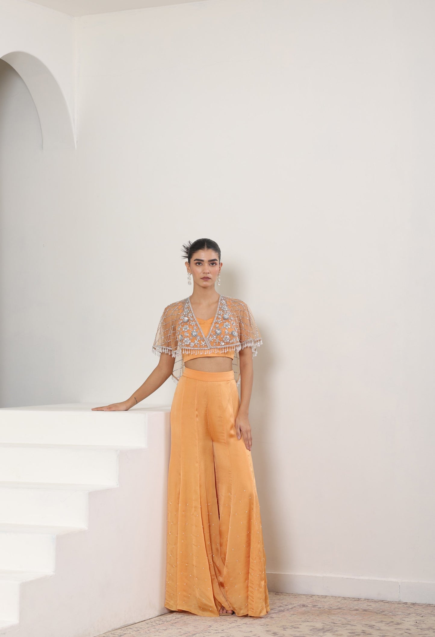 Woman in a peach outfit with intricate top and flowing pants standing on a white staircase. Orange satin organza sharara, embroidered cape set, festive ethnic outfit, elegant designer wear, premium traditional attire, Sunanta Madaan, near me, Mumbai, Delhi, Hyderabad, Bangalore, Pune
