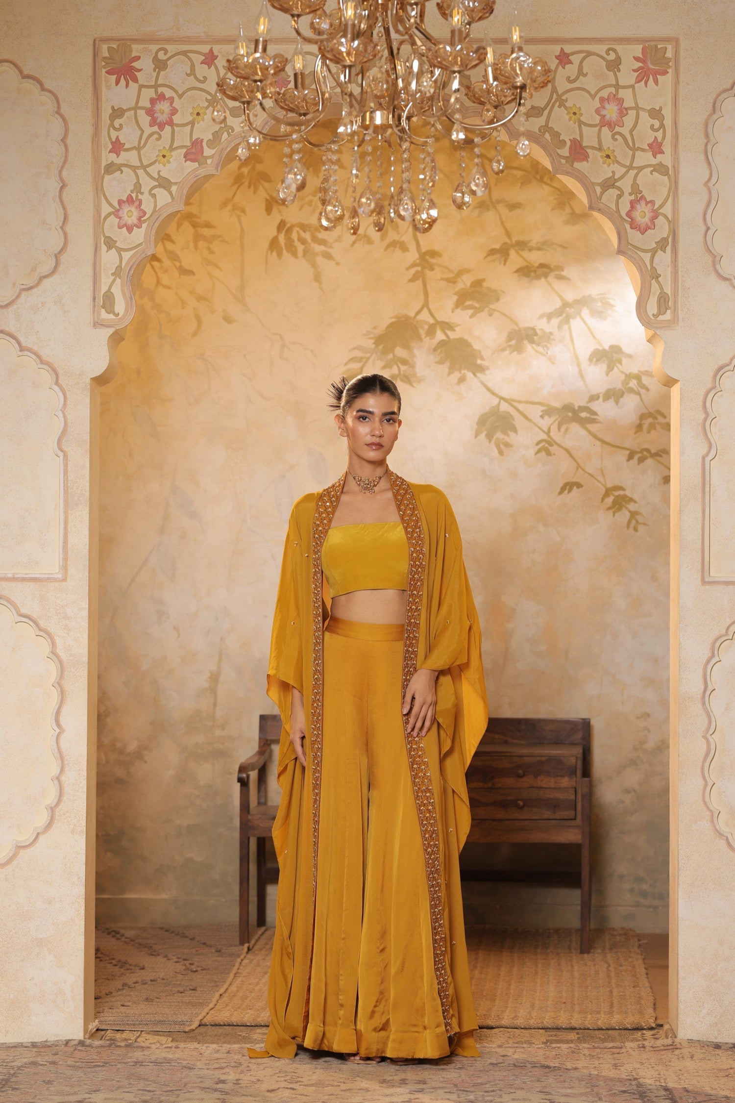 Woman in a mustard yellow outfit standing in an ornate room with a chandelier. Golden yellow sharara set, embroidered wedding outfit, designer festive wear, elegant ethnic ensemble, premium traditional attire, Sunanta Madaan