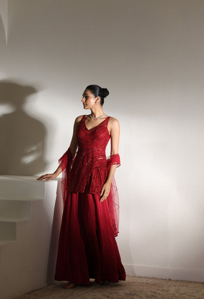 Woman in a red dress standing against a white wall with a soft shadow. Sunanta Madaan – Burgundy Satin Organza Sharara with Embroidered Corset, Wedding Wear, Elegant Festive Outfit, Boho Indo-Western Dress, Indo-Western Fusion Wear, Indo-Western Attire for Engagement, Indo-Western Outfit for Sangeet, Indo-Western Draped & Embroidered Skirt, Women’s Designer Party Wear, Delhi, Jodhpur, Hyderabad, Ahmedabad, Bangalore, near me