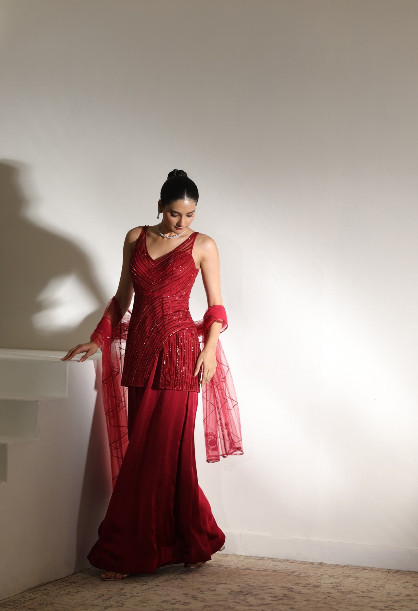 Woman in a red evening gown standing against a plain wall. Sunanta Madaan – Burgundy Satin Organza Sharara with Embroidered Corset, Wedding Wear, Elegant Festive Outfit, Boho Indo-Western Dress, Indo-Western Fusion Wear, Indo-Western Attire for Engagement, Indo-Western Outfit for Sangeet, Indo-Western Draped & Embroidered Skirt, Women’s Designer Party Wear, Delhi, Jodhpur, Hyderabad, Ahmedabad, Bangalore, near me