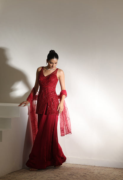 Woman in a red evening gown standing against a plain wall. Sunanta Madaan – Burgundy Satin Organza Sharara with Embroidered Corset, Wedding Wear, Elegant Festive Outfit, Boho Indo-Western Dress, Indo-Western Fusion Wear, Indo-Western Attire for Engagement, Indo-Western Outfit for Sangeet, Indo-Western Draped & Embroidered Skirt, Women’s Designer Party Wear, Delhi, Jodhpur, Hyderabad, Ahmedabad, Bangalore, near me