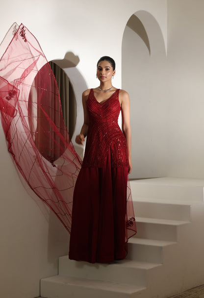 Woman in a red evening gown standing on white steps with a decorative red fabric in the background. Sunanta Madaan – Burgundy Satin Organza Sharara with Embroidered Corset, Wedding Wear, Elegant Festive Outfit, Boho Indo-Western Dress, Indo-Western Fusion Wear, Indo-Western Attire for Engagement, Indo-Western Outfit for Sangeet, Indo-Western Draped & Embroidered Skirt, Women’s Designer Party Wear, Delhi, Jodhpur, Hyderabad, Ahmedabad, Bangalore, near me