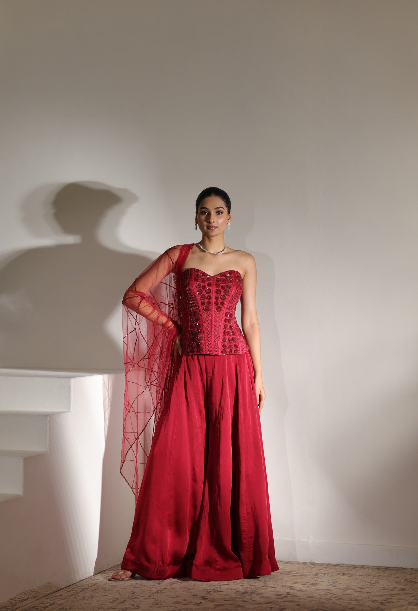 Woman wearing a red dress with a sheer overlay against a plain background; Sunanta Madaan – Maroon Satin Organza Embroidered Sharara Set, Premium Wedding Wear, Elegant Festive Outfit, Boho Indo-Western Dress, Indo-Western Fusion Wear, Indo-Western Attire for Engagement, Indo-Western Outfit for Sangeet, Indo-Western Draped & Embroidered Skirt, Women’s Designer Party Ensemble, Delhi, Jaipur, Hyderabad, Ahmedabad, Bangalore, near me
