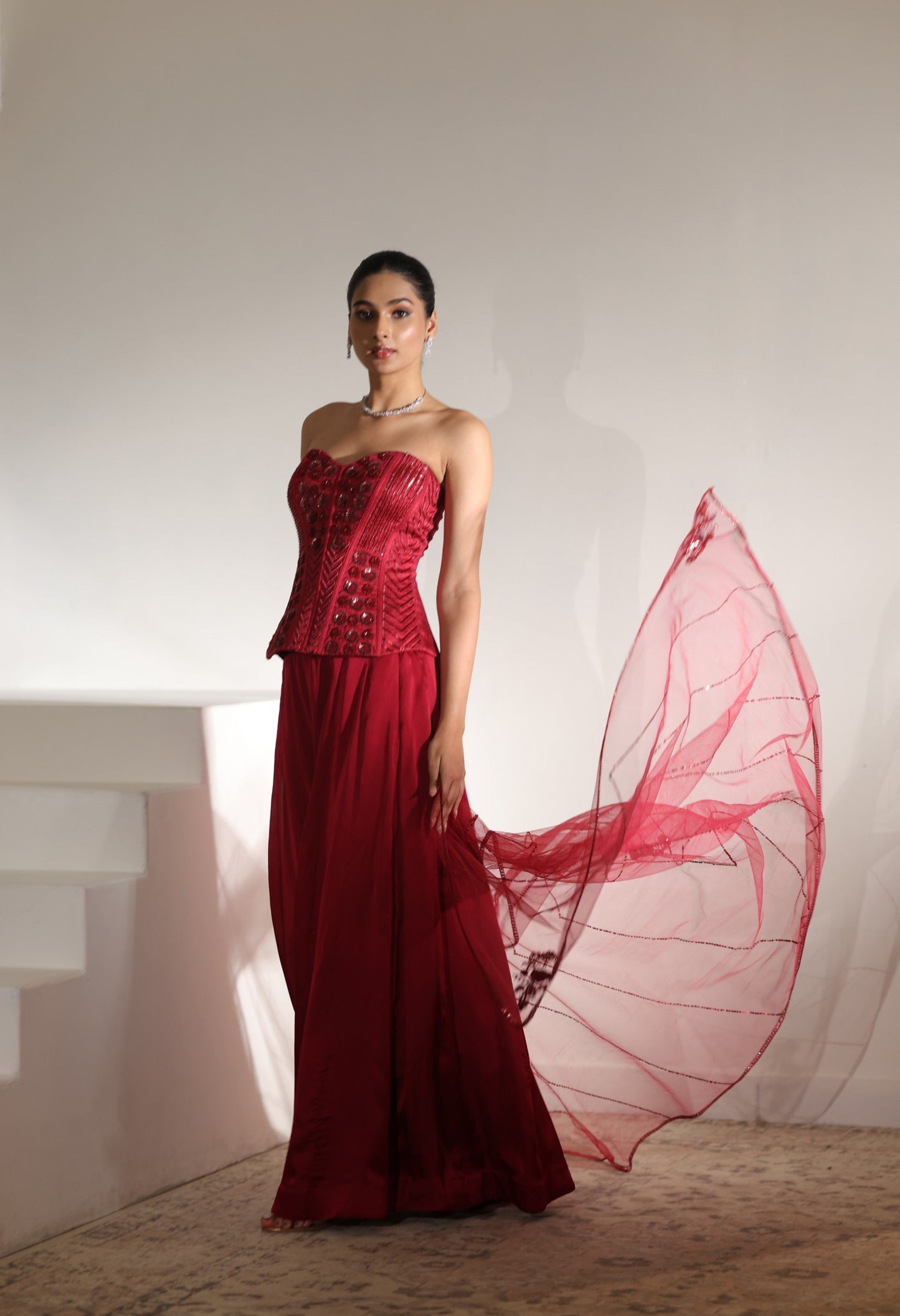 Woman wearing a red strapless gown with a sheer red overlay in a minimalistic setting; Sunanta Madaan – Maroon Satin Organza Embroidered Sharara Set, Premium Wedding Wear, Elegant Festive Outfit, Boho Indo-Western Dress, Indo-Western Fusion Wear, Indo-Western Attire for Engagement, Indo-Western Outfit for Sangeet, Indo-Western Draped & Embroidered Skirt, Women’s Designer Party Ensemble, Delhi, Jaipur, Hyderabad, Ahmedabad, Bangalore, near me