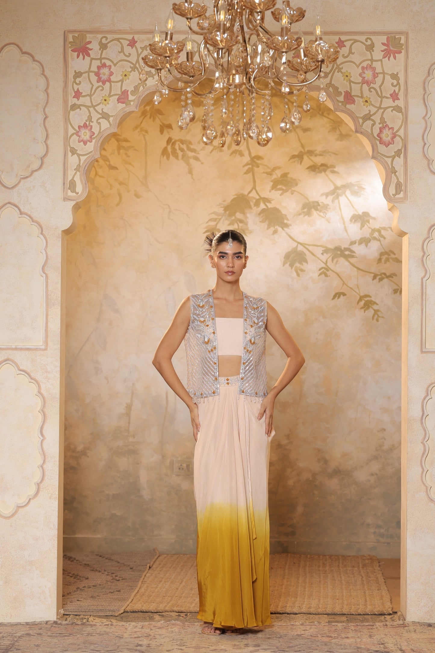 Woman in a traditional outfit with a decorative background. Ivory yellow draped skirt set, handcrafted crepe outfit, luxury evening wear, elegant festive attire, designer ethnic ensemble, Sunanta Madaan, near me, Mumbai, Delhi, Hyderabad, Bangalore, Pune