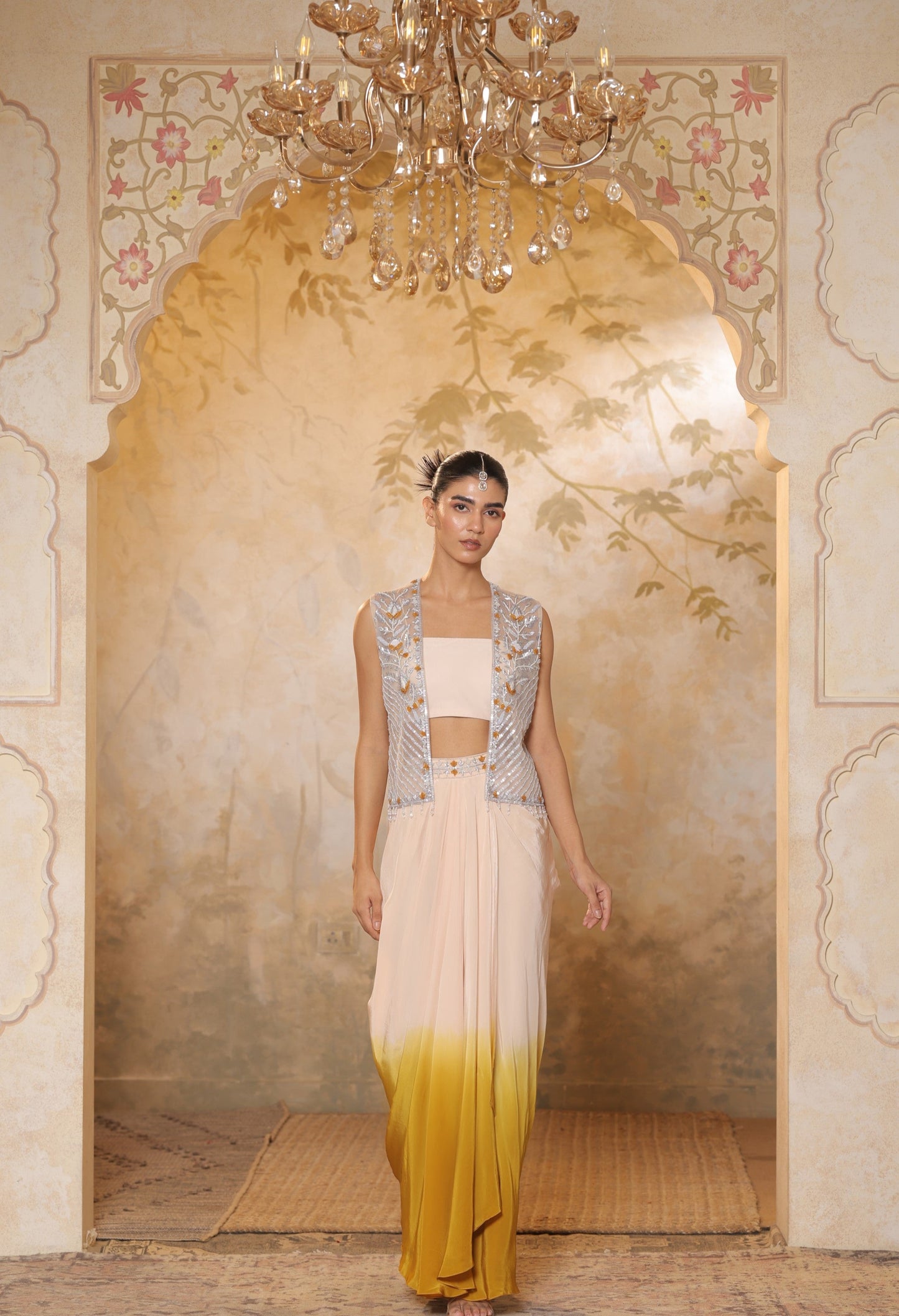 Woman in a traditional outfit standing in an ornate room with a chandelier. Ivory yellow draped skirt set, handcrafted crepe outfit, luxury evening wear, elegant festive attire, designer ethnic ensemble, Sunanta Madaan, near me, Mumbai, Delhi, Hyderabad, Bangalore, Pune