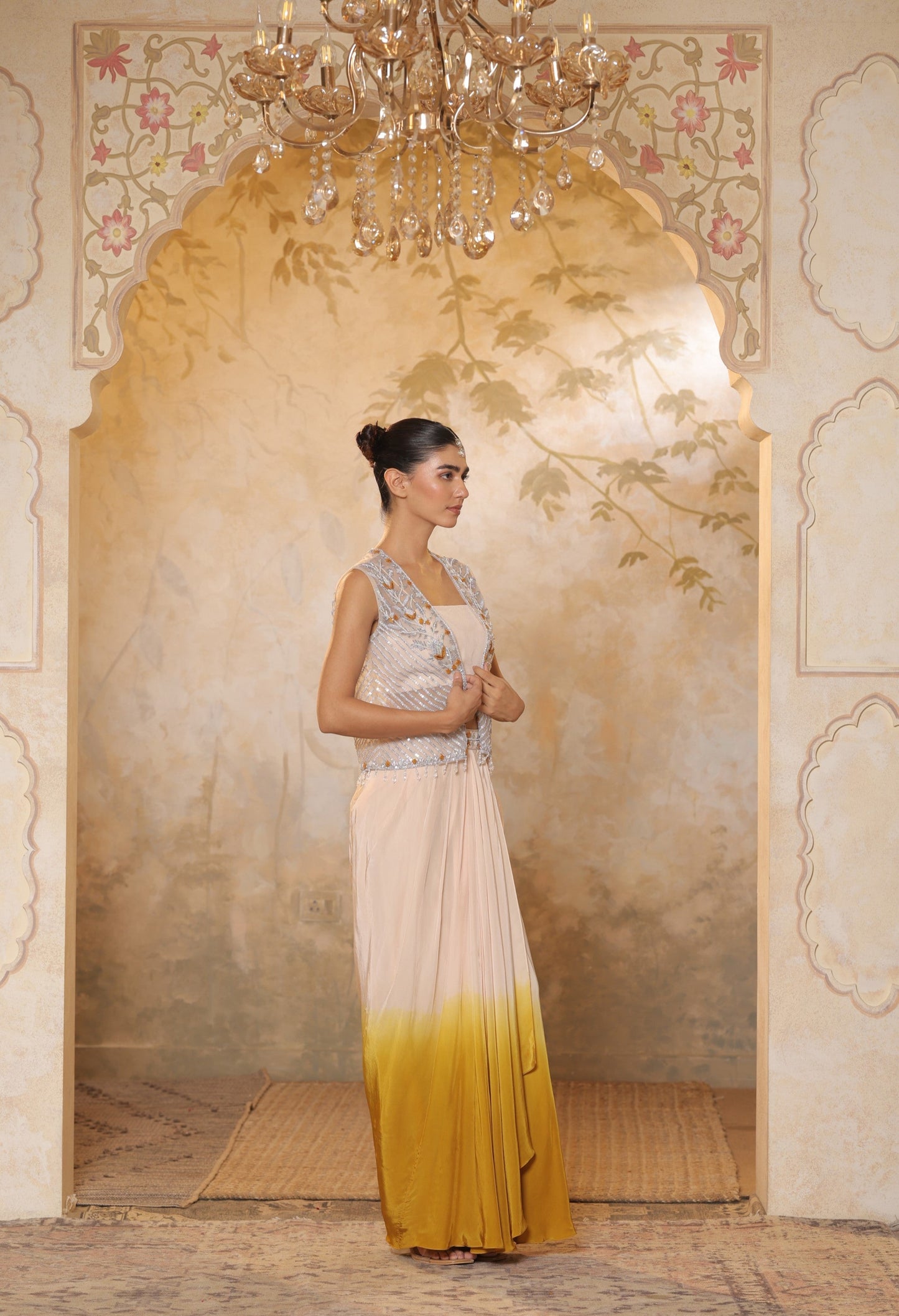 Woman in a traditional outfit standing in an ornate room with a chandelier. Ivory yellow draped skirt set, handcrafted crepe outfit, luxury evening wear, elegant festive attire, designer ethnic ensemble, Sunanta Madaan, near me, Mumbai, Delhi, Hyderabad, Bangalore, Pune