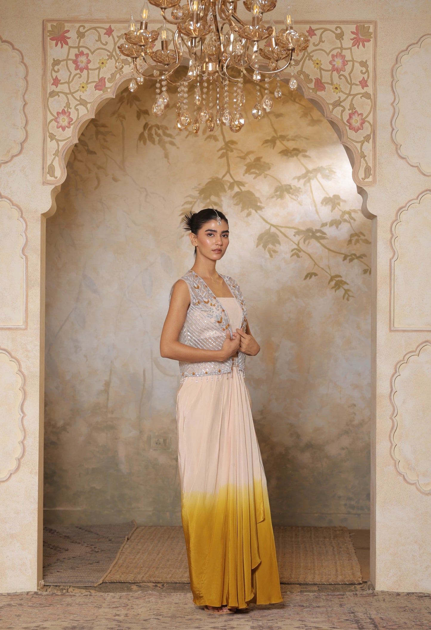 Woman in a traditional outfit standing in an ornate room with a chandelier. Ivory yellow draped skirt set, handcrafted crepe outfit, luxury evening wear, elegant festive attire, designer ethnic ensemble, Sunanta Madaan, near me, Mumbai, Delhi, Hyderabad, Bangalore, Pune