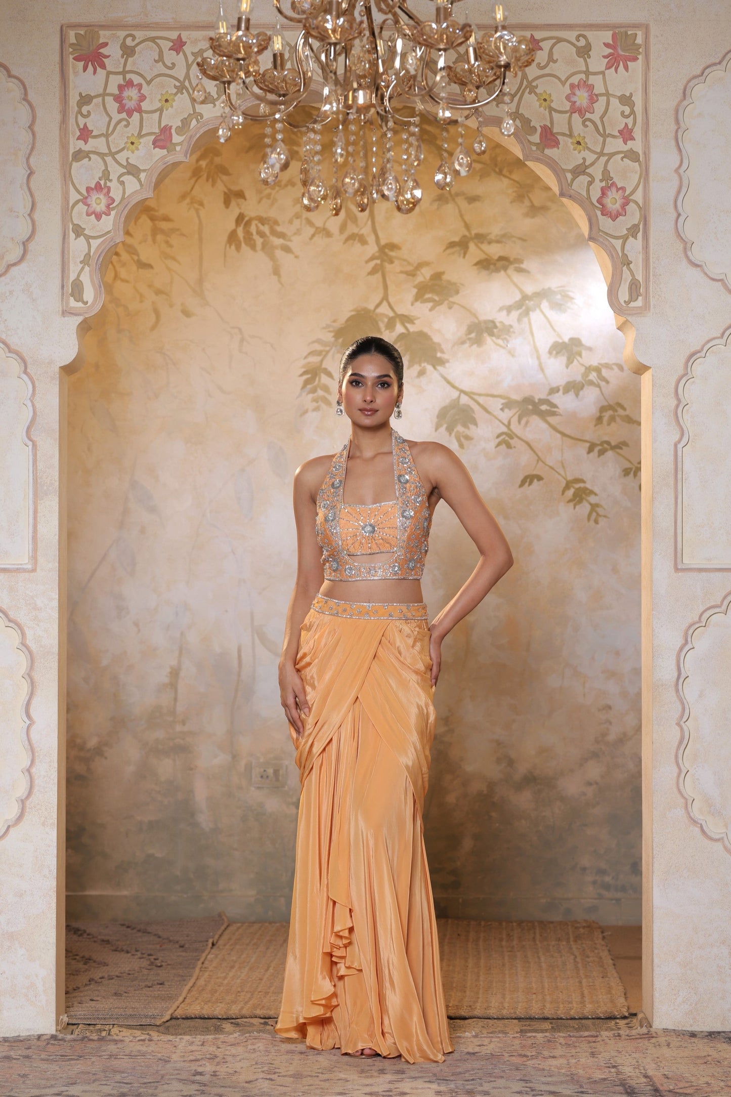Woman in a peach saree with silver blouse standing in a decorated room with floral arches and chandelier. Orange viscose crepe skirt set, premium festive outfit, elegant ethnic wear, designer traditional attire, handcrafted party ensemble, Sunanta Madaan, near me, Mumbai, Delhi, Hyderabad, Bangalore, Pune