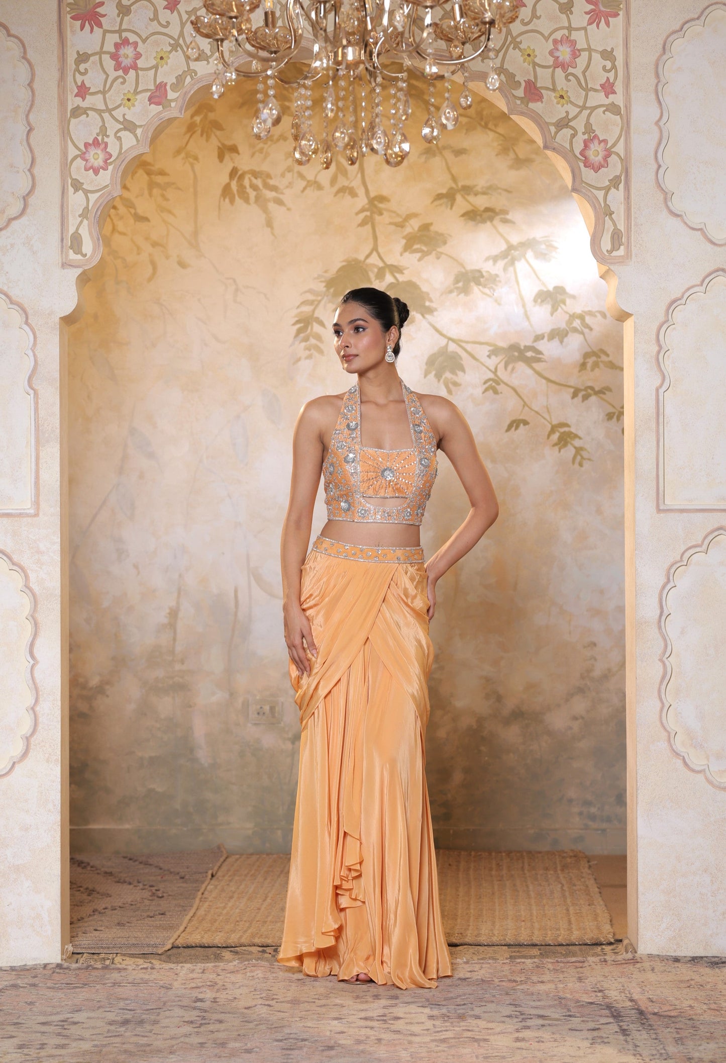 Woman in a peach saree with intricate design standing in an ornate room with a chandelier. Orange viscose crepe skirt set, premium festive outfit, elegant ethnic wear, designer traditional attire, handcrafted party ensemble, Sunanta Madaan, near me, Mumbai, Delhi, Hyderabad, Bangalore, Pune