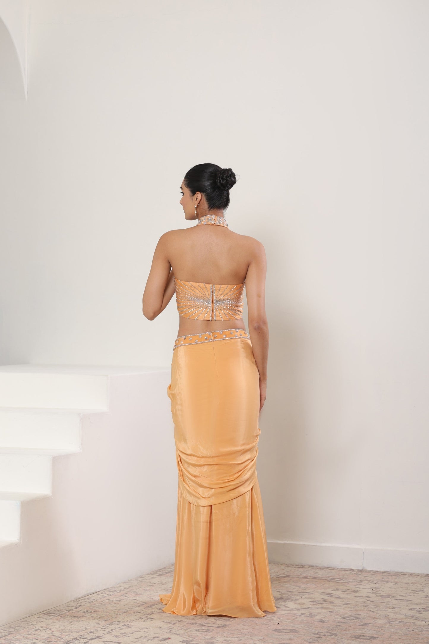 Woman in a strapless, long, orange evening gown standing against a white wall. Orange viscose crepe skirt set, premium festive outfit, elegant ethnic wear, designer traditional attire, handcrafted party ensemble, Sunanta Madaan, near me, Mumbai, Delhi, Hyderabad, Bangalore, Pune