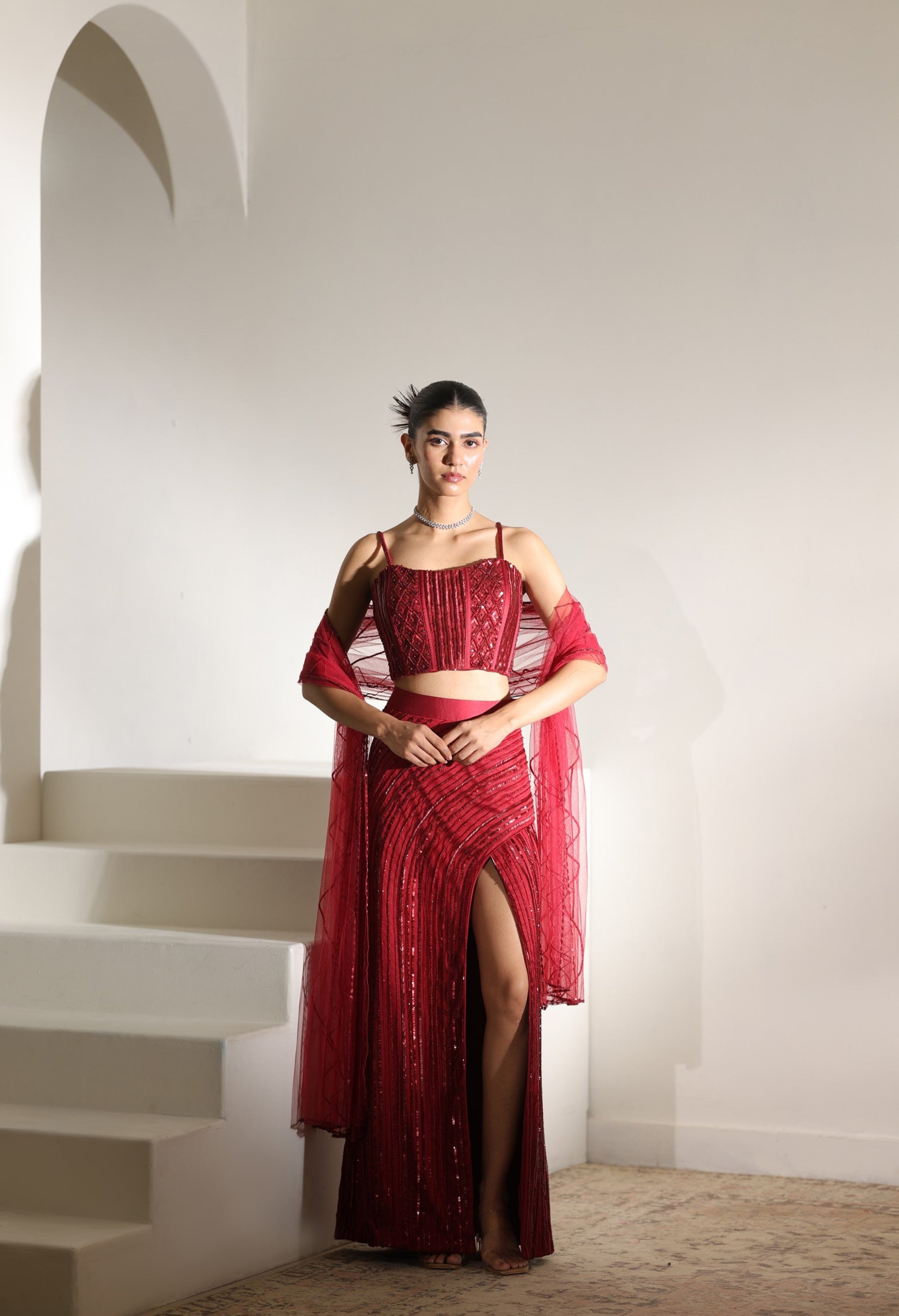 Woman in a red dress standing on a staircase with a white wall background; Sunanta Madaan – Burgundy Satin Embroidered Western Skirt Set, Handcrafted Festive Wear, Elegant Party Outfit, Boho Indo-Western Dress, Indo-Western Fusion Wear, Indo-Western Attire for Engagement, Indo-Western Outfit for Sangeet, Indo-Western Draped & Embroidered Skirt, Women’s Designer Festive Ensemble, Mumbai, Jaipur, Hyderabad, Ahmedabad, Pune, near me