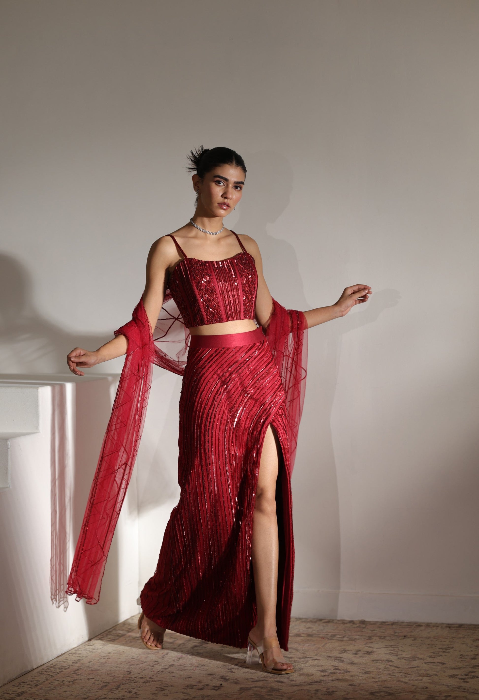 Woman in a red dress with a slit standing against a plain wall. Sunanta Madaan – Burgundy Satin Embroidered Western Skirt Set, Handcrafted Festive Wear, Elegant Party Outfit, Boho Indo-Western Dress, Indo-Western Fusion Wear, Indo-Western Attire for Engagement, Indo-Western Outfit for Sangeet, Indo-Western Draped & Embroidered Skirt, Women’s Designer Festive Ensemble, Mumbai, Jaipur, Hyderabad, Ahmedabad, Pune, near me
