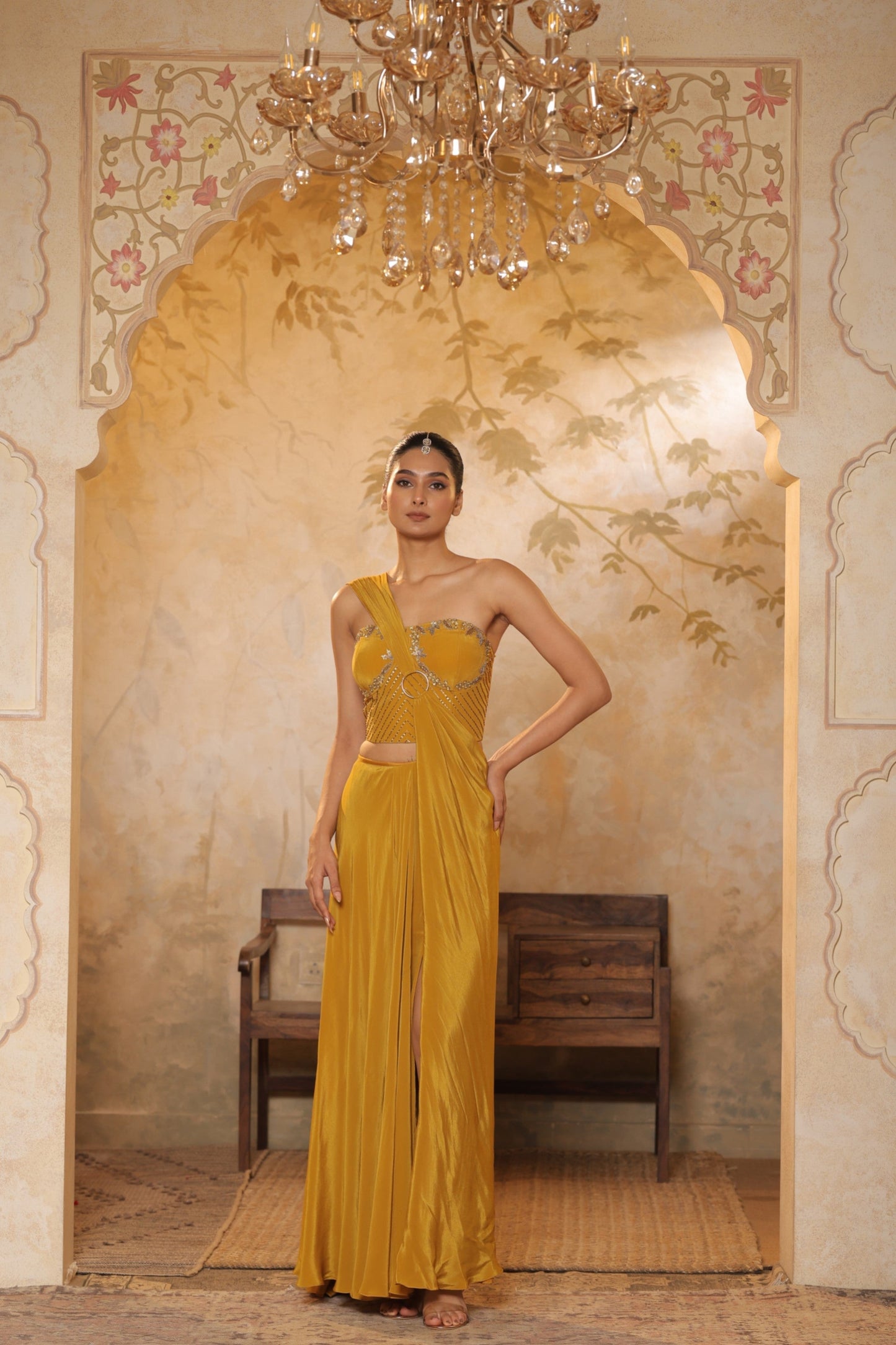 Woman in a yellow evening gown standing in an ornate room with a chandelier. Golden yellow crepe saree, embroidered haldi saree, premium festive wear, elegant ethnic outfit, designer traditional saree, Sunanta Madaan