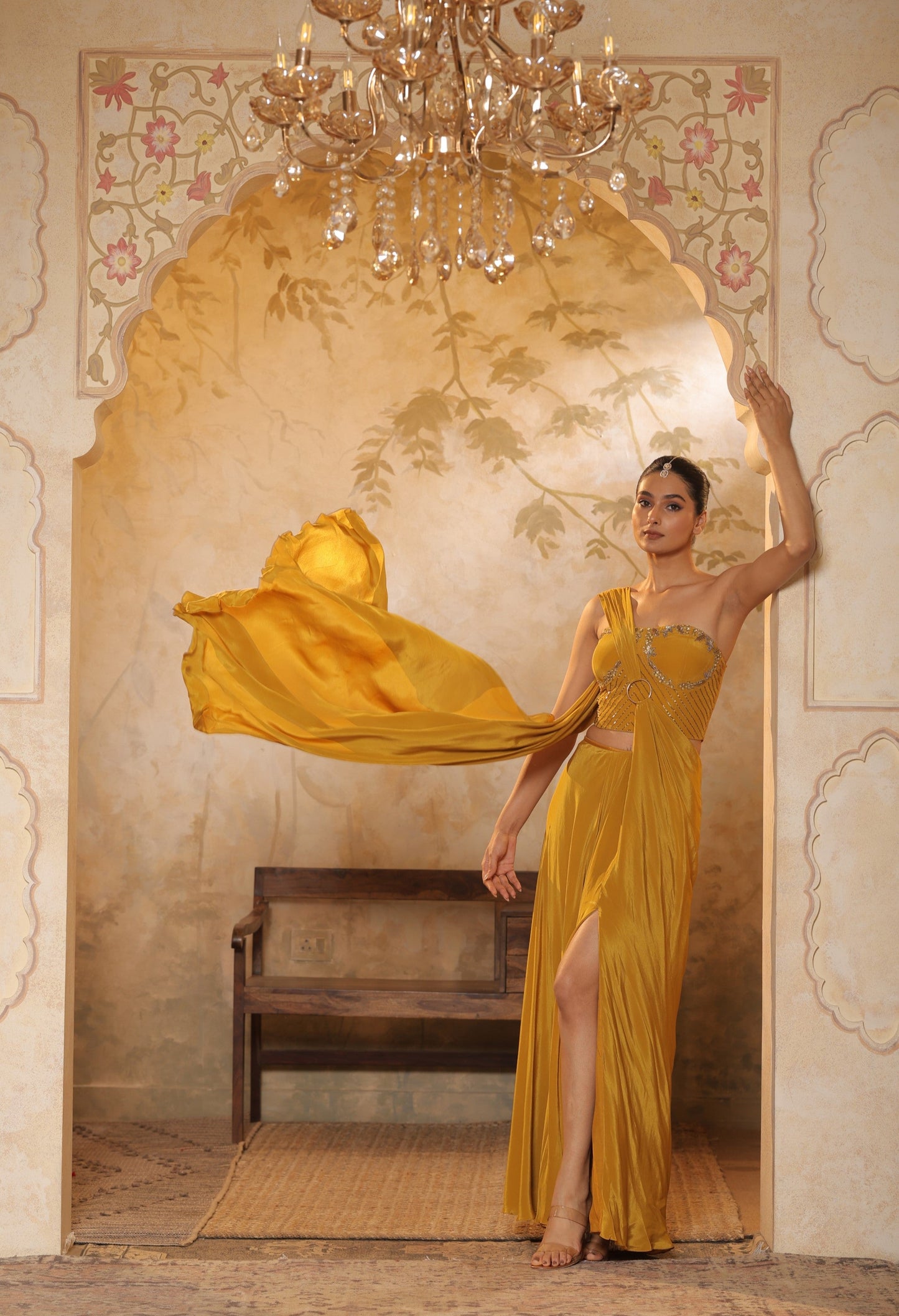 Woman in a yellow dress standing in an ornate room with a chandelier. Golden yellow crepe saree, embroidered haldi saree, premium festive wear, elegant ethnic outfit, designer traditional saree, Sunanta Madaan