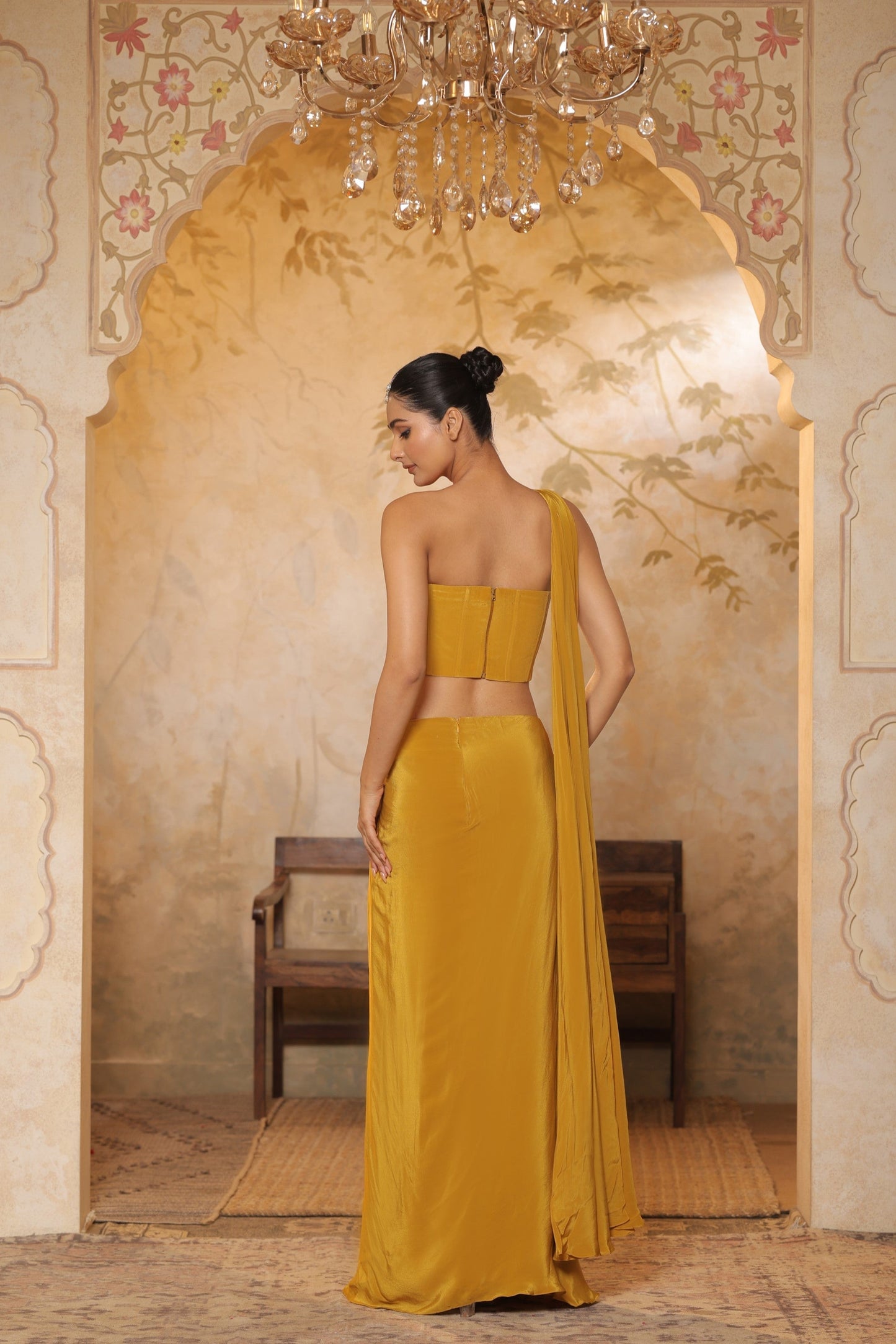 Woman in a mustard yellow saree standing in an ornate room with a chandelier. Golden yellow crepe saree, embroidered haldi saree, premium festive wear, elegant ethnic outfit, designer traditional saree, Sunanta Madaan