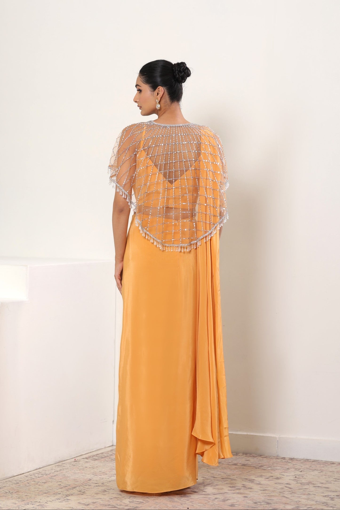 Woman wearing a mustard yellow saree dress with a textured overlay in a minimal indoor setting; designer reception saree, elegant festive outfit, Orange crepe saree, embroidered cape saree, premium evening wear, Sunanta Madaan