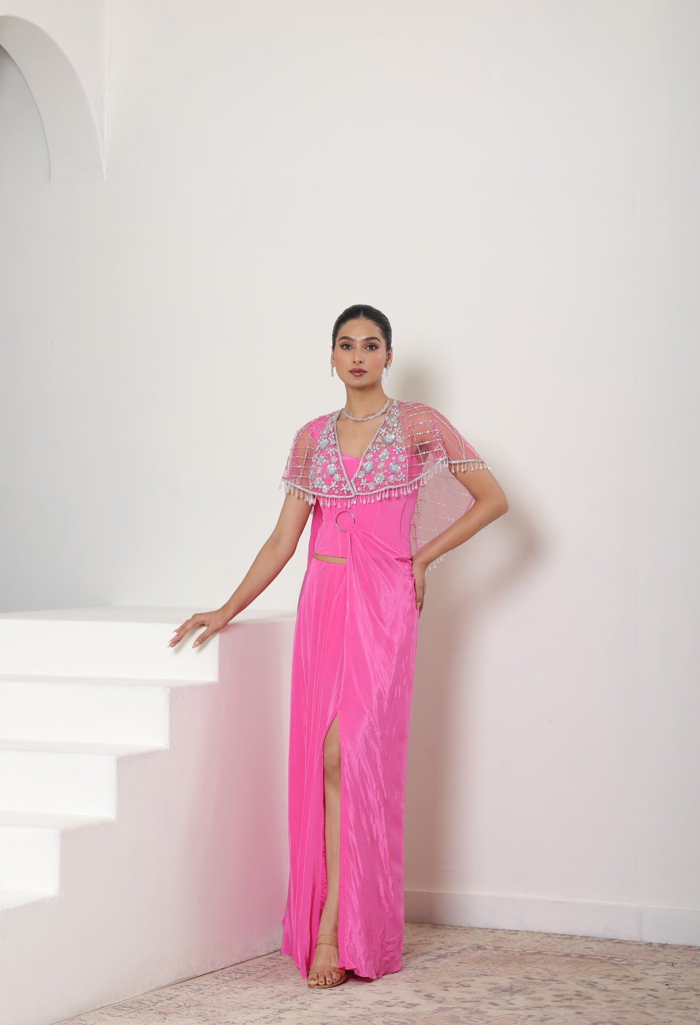 Woman in a pink evening gown with floral embellishments standing on a staircase. Pink crepe saree, embroidered cape saree,elegant bridal wear, festive ethnic outfit, Sunanta Madaan