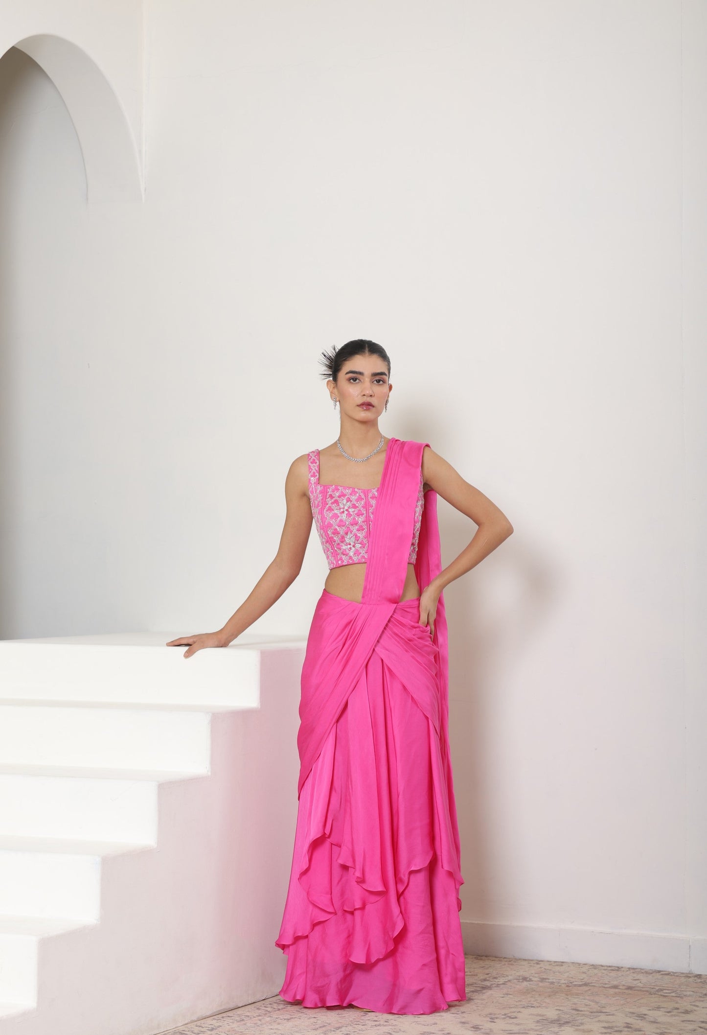 Woman in a pink saree standing on white steps with a white wall background; Pink satin organza saree, premium sangeet saree, embroidered designer saree, elegant festive wear, party ready saree, Sunanta Madaan