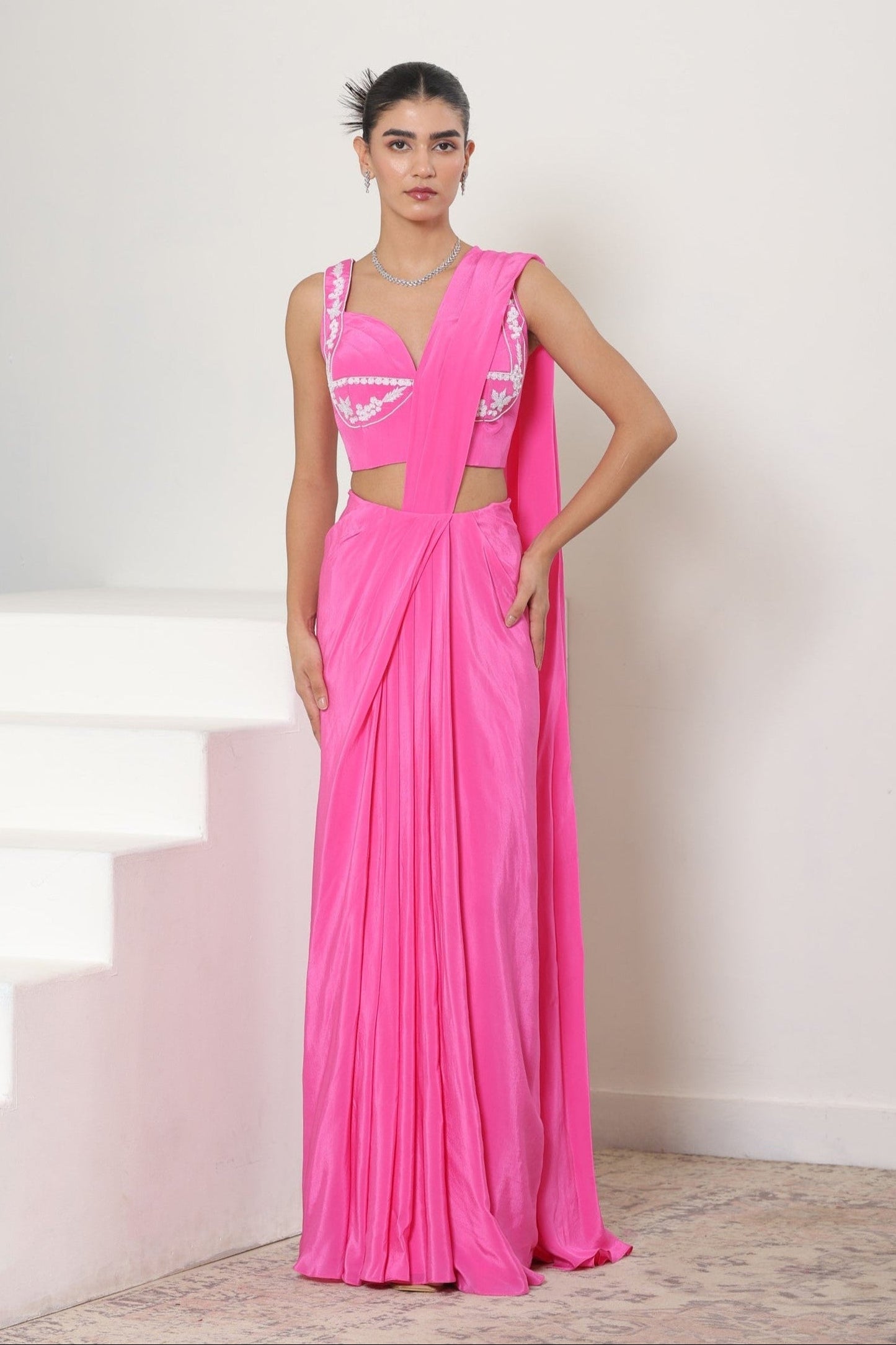 Woman wearing a pink saree with a white blouse, standing on a staircase. Pink crepe saree, embroidered cocktail saree, premium party wear saree, designer festive outfit, elegant evening saree, Sunanta Madaan