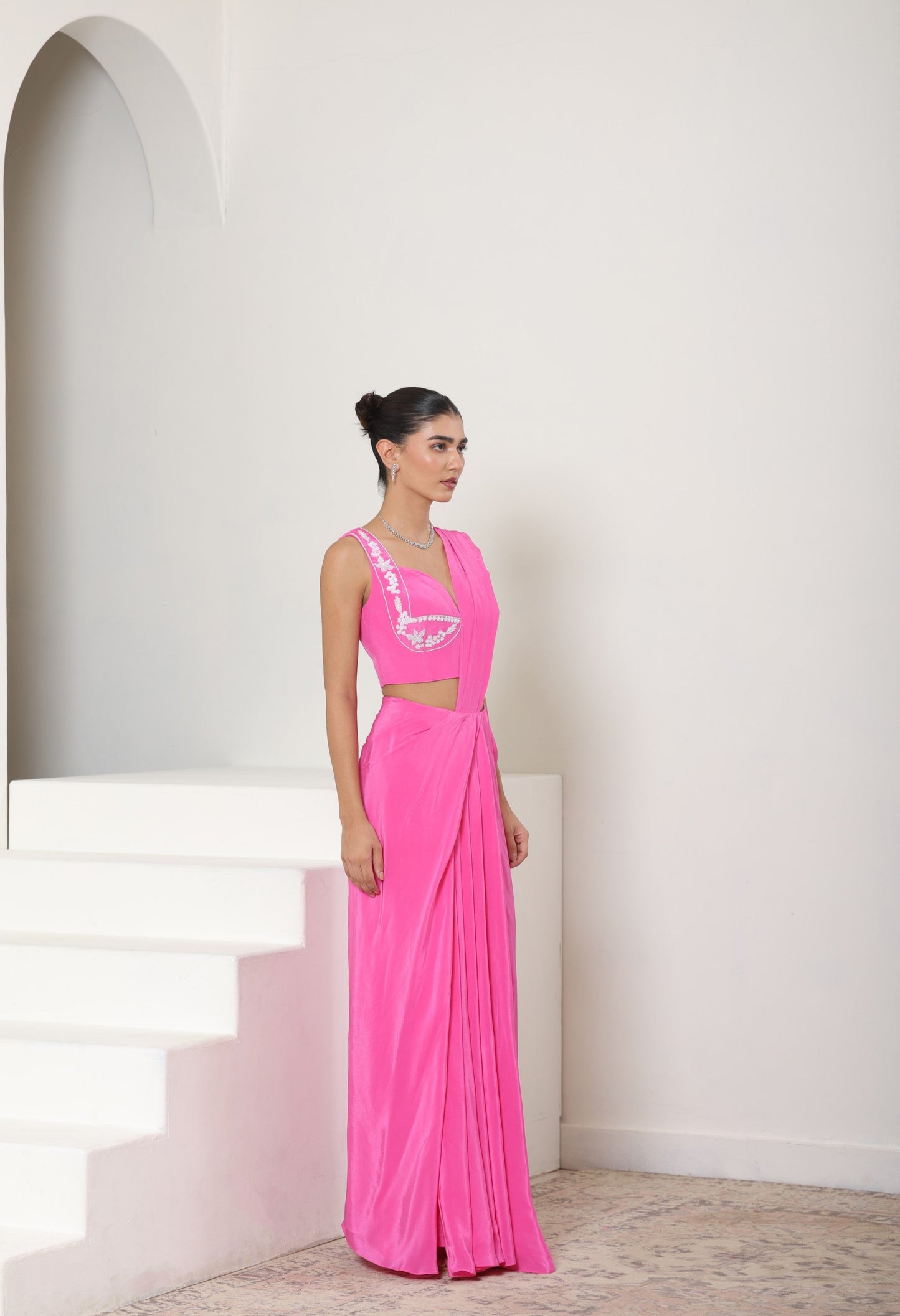 Woman in a pink dress standing on white stairs with a white wall background; Pink crepe saree, embroidered cocktail saree, premium party wear saree, designer festive outfit, elegant evening saree, Sunanta Madaan