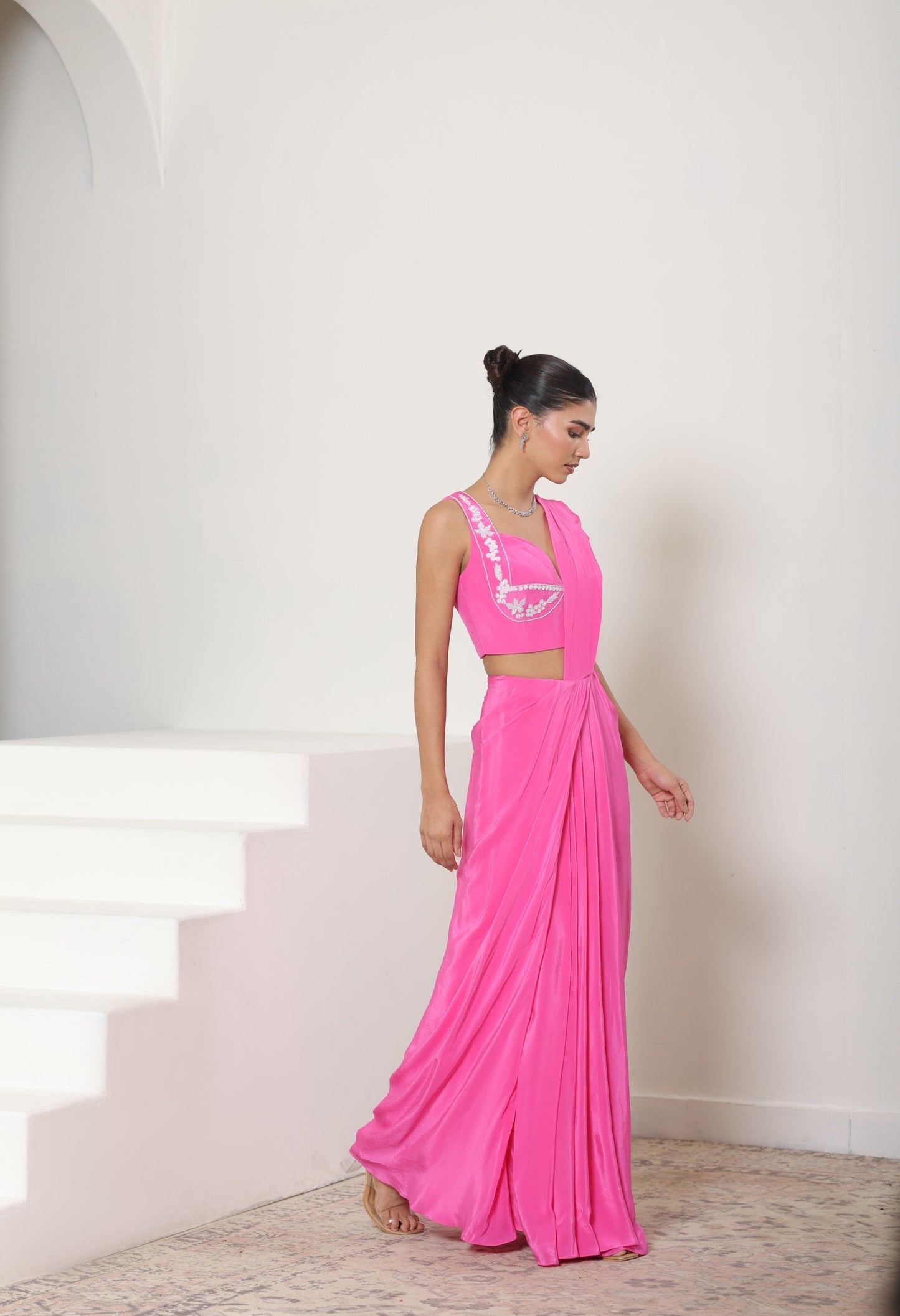Woman in a pink saree standing on a staircase with a white wall background; Pink crepe saree, embroidered cocktail saree, premium party wear saree, designer festive outfit, elegant evening saree, Sunanta Madaan