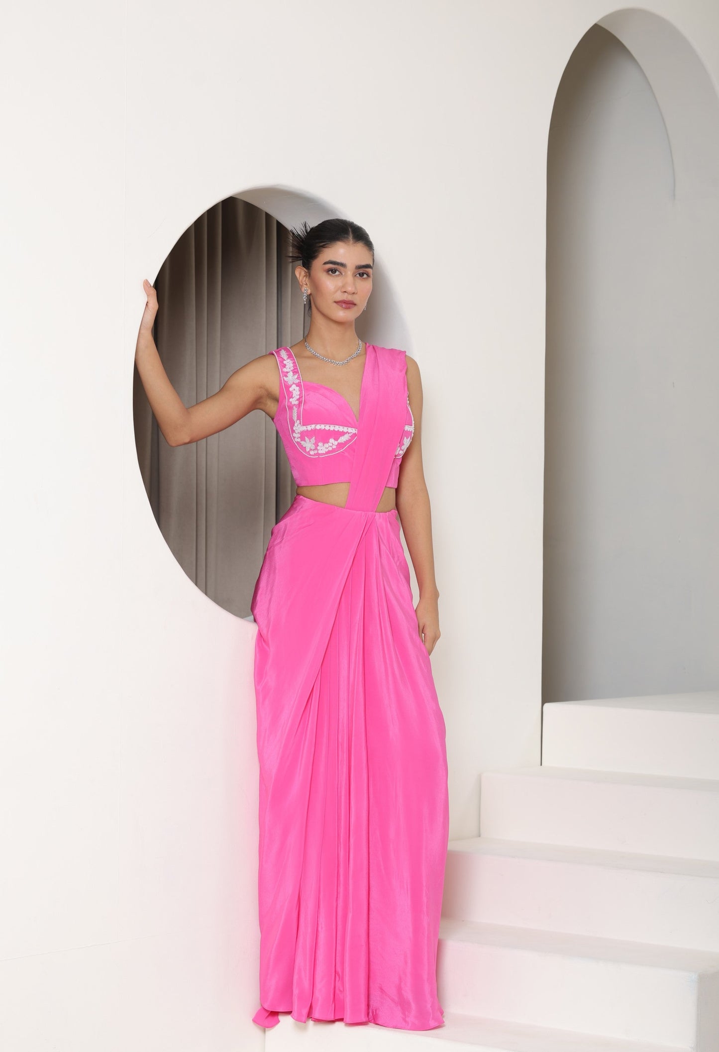 Woman wearing a bright pink dress with cut-out design in a minimalistic setting. Pink crepe saree, embroidered cocktail saree, premium party wear saree, designer festive outfit, elegant evening saree, Sunanta Madaan