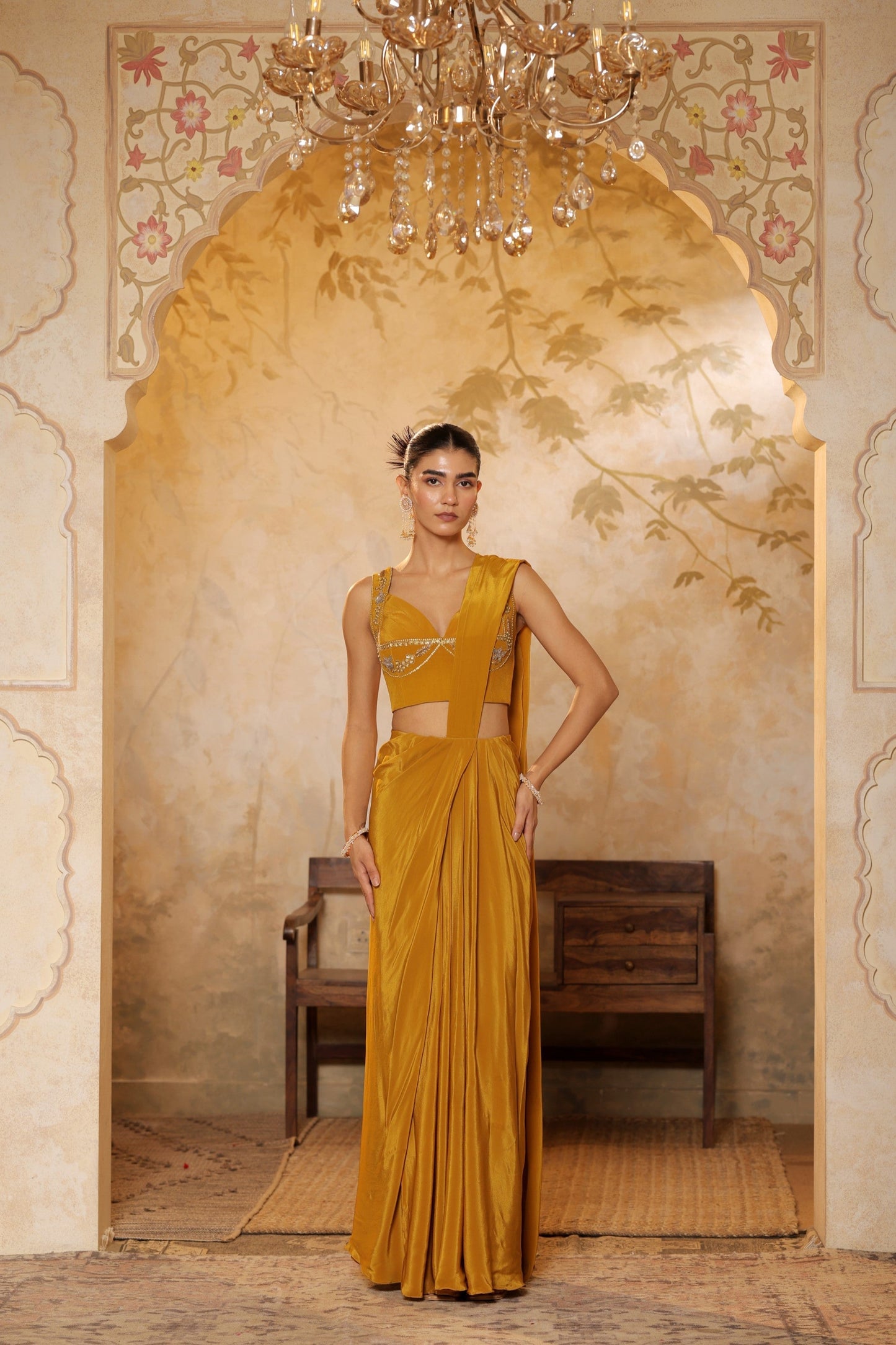 Woman in a yellow saree standing in an ornate room with a chandelier. Golden yellow crepe saree, haldi ceremony saree, embroidered festive saree, designer ethnic wear, elegant haldi outfit, Sunanta Madaan